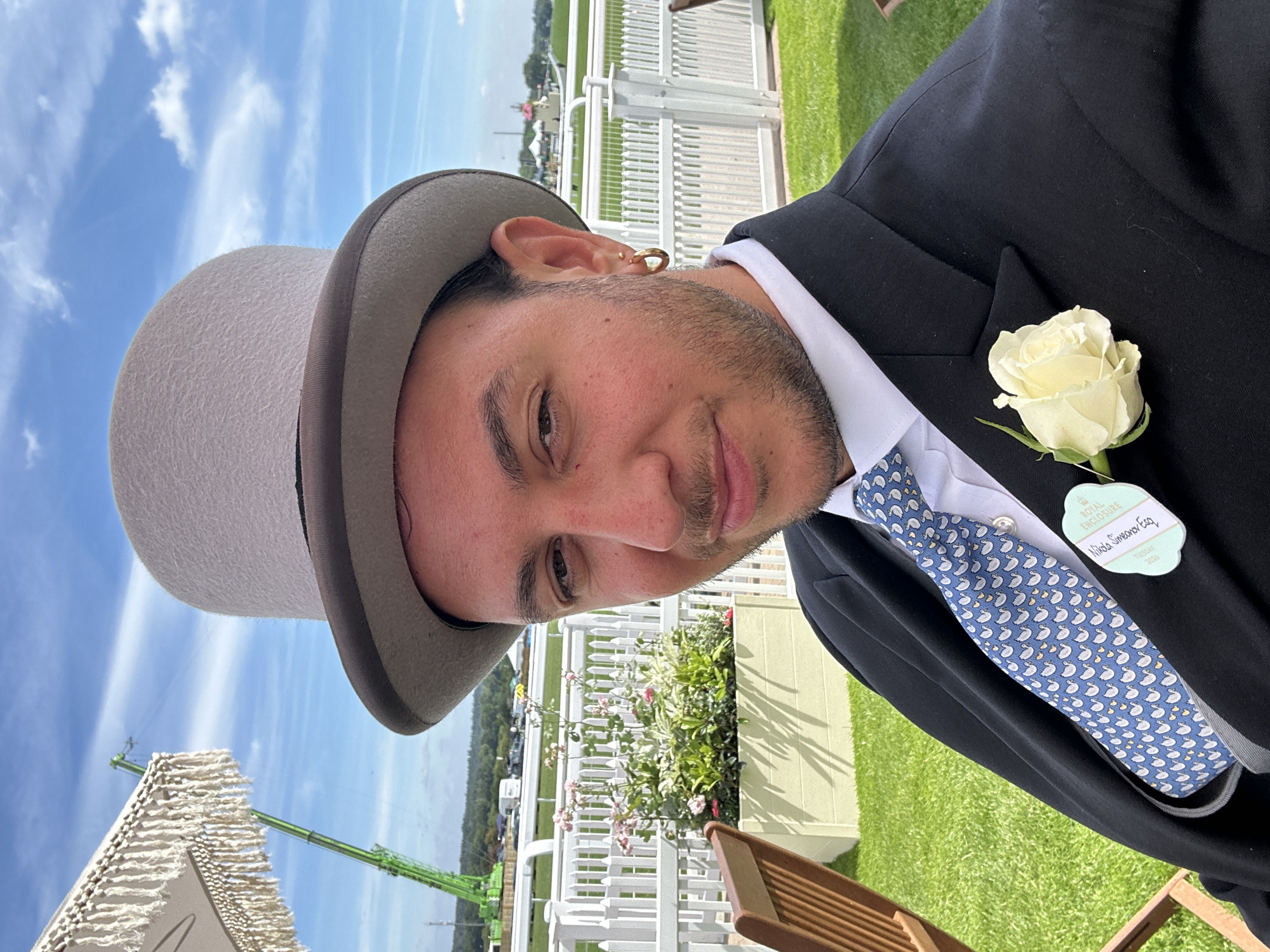 A man wearing a grey top hat, black suit, and a blue tie with a duck pattern poses for a selfie outdoors. He has a white rose pinned to his lapel. In the background, a white picket fence runs along a green lawn, with a flower planter and chairs visible. Beyond the fence is a racetrack, a crane, and tents. The sky is blue with scattered clouds. A badge attached to his jacket reads "Royal Enclosure Tuesday". The scene is likely at Ascot Racecourse in Ascot, United Kingdom, a venue known for its horse racing events and strict dress code.