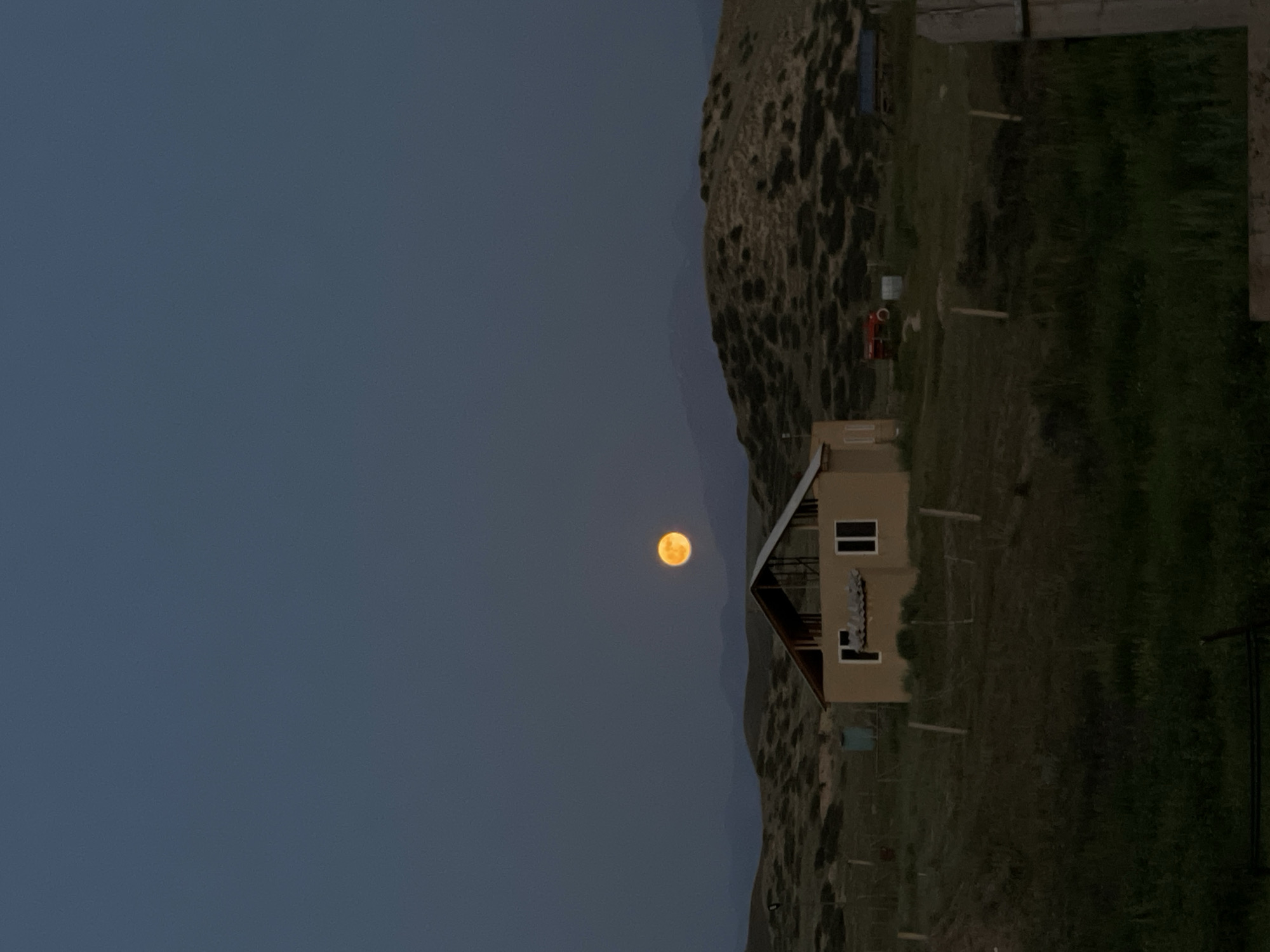 The image shows a rural scene at dusk, with a full moon prominently displayed in the dark blue sky. The moon is a bright, orange-yellow hue, appearing large and luminous against the fading light.
In the mid-ground, a small, beige house with a steep, dark roof is visible. It has a simple design with windows and what appears to be an open-air porch or balcony. The house is situated on a grassy hillside.
The landscape features rolling hills and sparse vegetation, characteristic of a mountainous or arid region. There are some wooden posts and fencing scattered across the terrain, suggesting it might be used for livestock or property demarcation. A small red tractor or farm implement is parked near the house. The overall setting is remote and tranquil.
The time of day is clearly evening, with the sky transitioning from twilight blue to darkness, and the moon being the primary source of illumination aside from ambient light. The weather appears clear, allowing for an unobstructed view of the moon and the landscape. The location is identified as Kara-Oi, Kyrgyzstan, which aligns with the rugged, hilly terrain. No people are visible in the image, and there is no visible text. The scene evokes a sense of peace and natural beauty, with the full moon casting an ethereal glow over the quiet landscape.