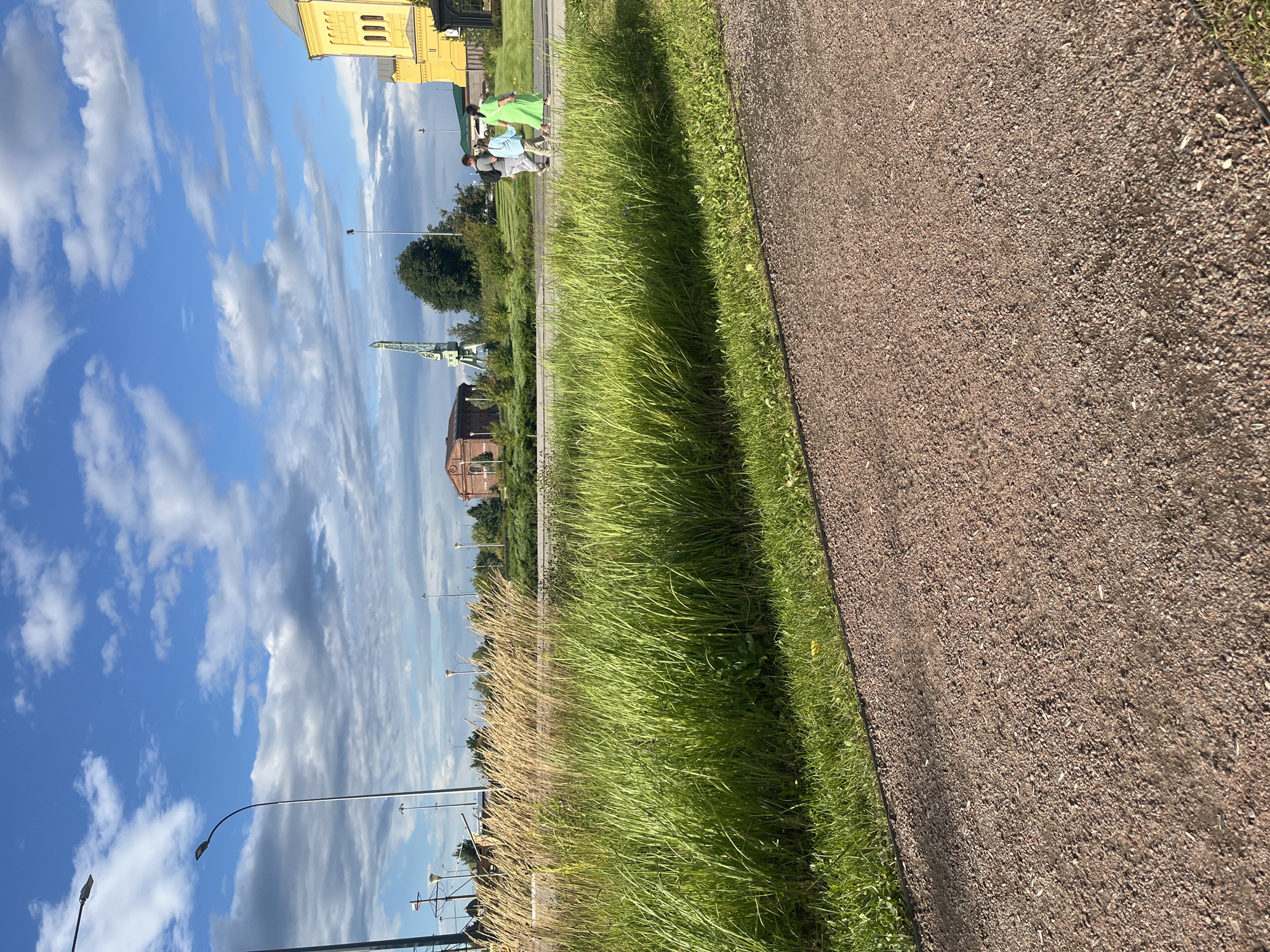 The image captures a scenic outdoor view, likely on a sunny day with scattered clouds in a bright blue sky. In the foreground, a gravel path runs diagonally across the frame, bordered by lush green grass and taller, dry grass. Further back, a road is visible with several streetlights. Across the road, there's a vibrant green industrial crane and a building with a red brick facade. To the right, a bright yellow building with arched windows stands prominently, possibly a church or historical structure. Three people are walking on the road in the middle ground, appearing to be casually strolling. While the image is generally clear, the specific location in Nizhny Novgorod, Russia, is not explicitly identifiable from the visual elements alone.