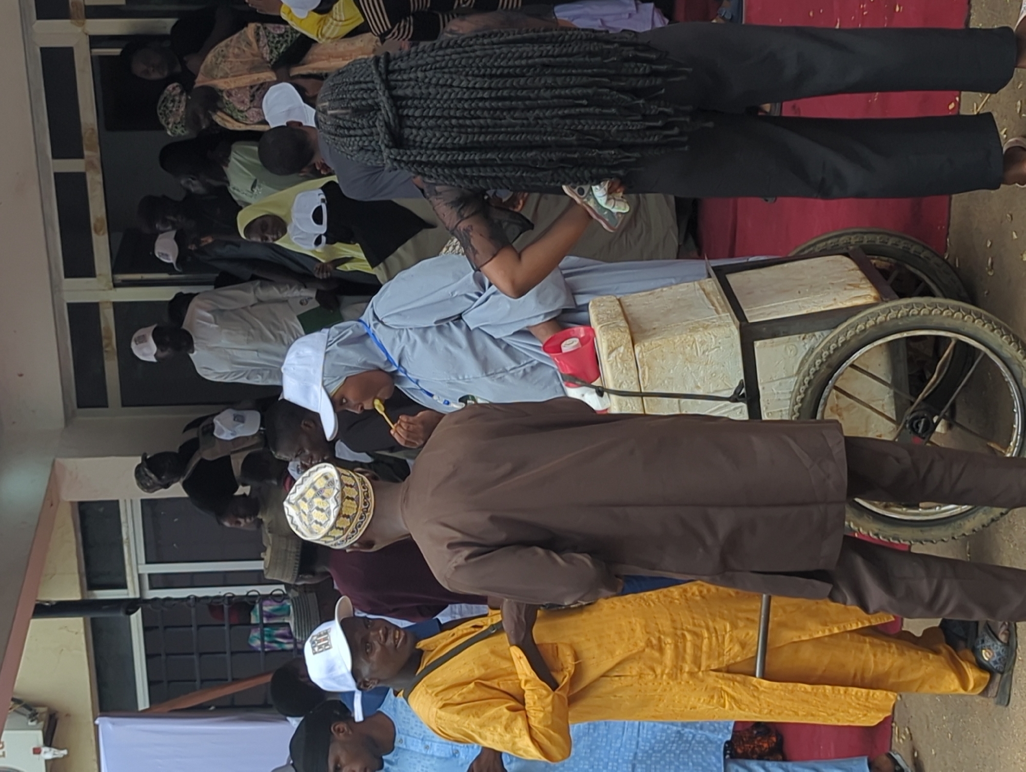 The image depicts a gathering of people in Ungwan Liman, Nigeria. Several individuals are dressed in traditional attire, including a man in a yellow robe and others wearing white caps. One person is eating with a spoon, while another leans over a cart-like structure with wheels. The background shows a building with windows and a red rug on the ground. Some people are observing from the building. Braided hair is visible on one individual.