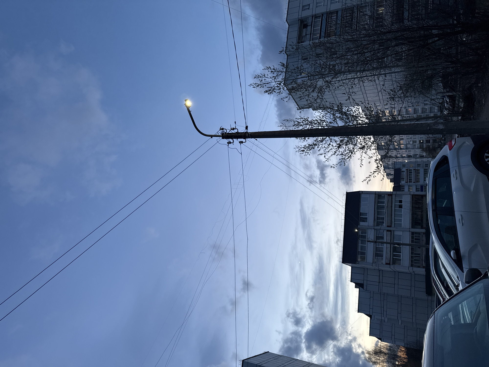 The image, rotated counter-clockwise, depicts an urban street scene at dusk or early evening in Chaikovsky, Russia. The sky dominates the upper and left portions, displaying a soft blue hue mixed with scattered grey clouds, some catching a subtle, fading light.
A prominent utility pole stands centrally, supporting a brightly illuminated street light that casts a distinct yellow-white glow. Numerous dark power lines and communication wires crisscross the sky, emanating from the pole and extending across the frame, forming complex linear patterns.
In the background and midground, several multi-story residential buildings, consistent with typical Russian urban architecture, are visible. These structures appear in shades of grey, with numerous windows. Bare tree branches, indicative of a dormant season like late autumn or early spring, are visible near the buildings and around the utility pole.
On the right side of the image, parts of two parked cars are visible. A white car, possibly a sedan or small SUV, shows its rear and side, while a darker vehicle is partially seen in the immediate foreground.
No people are visible, and no specific activity is taking place. The scene evokes a calm, quiet atmosphere of an urban residential area transitioning into night. There is no visible text.