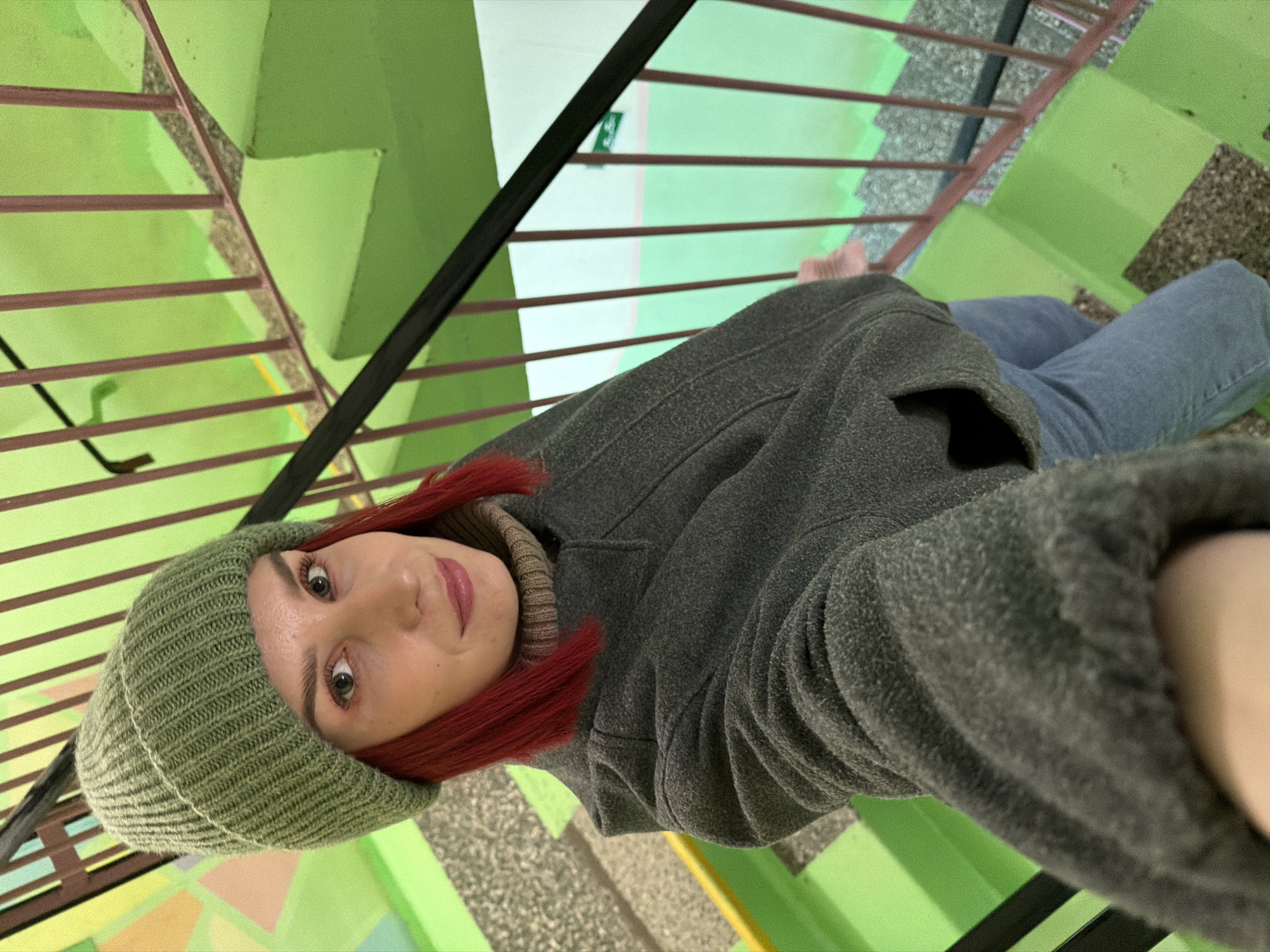 A medium-close-up, rotated selfie shows a young woman in a brightly colored indoor stairwell. The woman has light eyes and red hair, partially covered by a ribbed, olive green beanie. She is wearing a dark gray fleece jacket or coat over a light brown/beige ribbed turtleneck and blue jeans. Her expression is neutral to slightly smiling as she looks directly at the camera.
The background features vivid lime green walls and stairs, with the stair treads or risers appearing to be made of a mottled, dark aggregate material (possibly concrete or terrazzo). A railing with multiple horizontal bars, reddish-brown in color, separates the woman from the stairwell void. Another section of the railing, darker and possibly the one she's holding, is visible closer to the viewer.
The image perspective is rotated approximately 90 degrees clockwise, suggesting a dynamic pose or angle. In the distance, other sections of the green stairwell are visible, along with a small green sign featuring a white symbol (possibly an arrow or person pictogram) on a distant wall. The lighting appears to be artificial or diffused, illuminating the vibrant green surfaces. The setting suggests an institutional building or apartment complex in Chaikovsky, Russia.