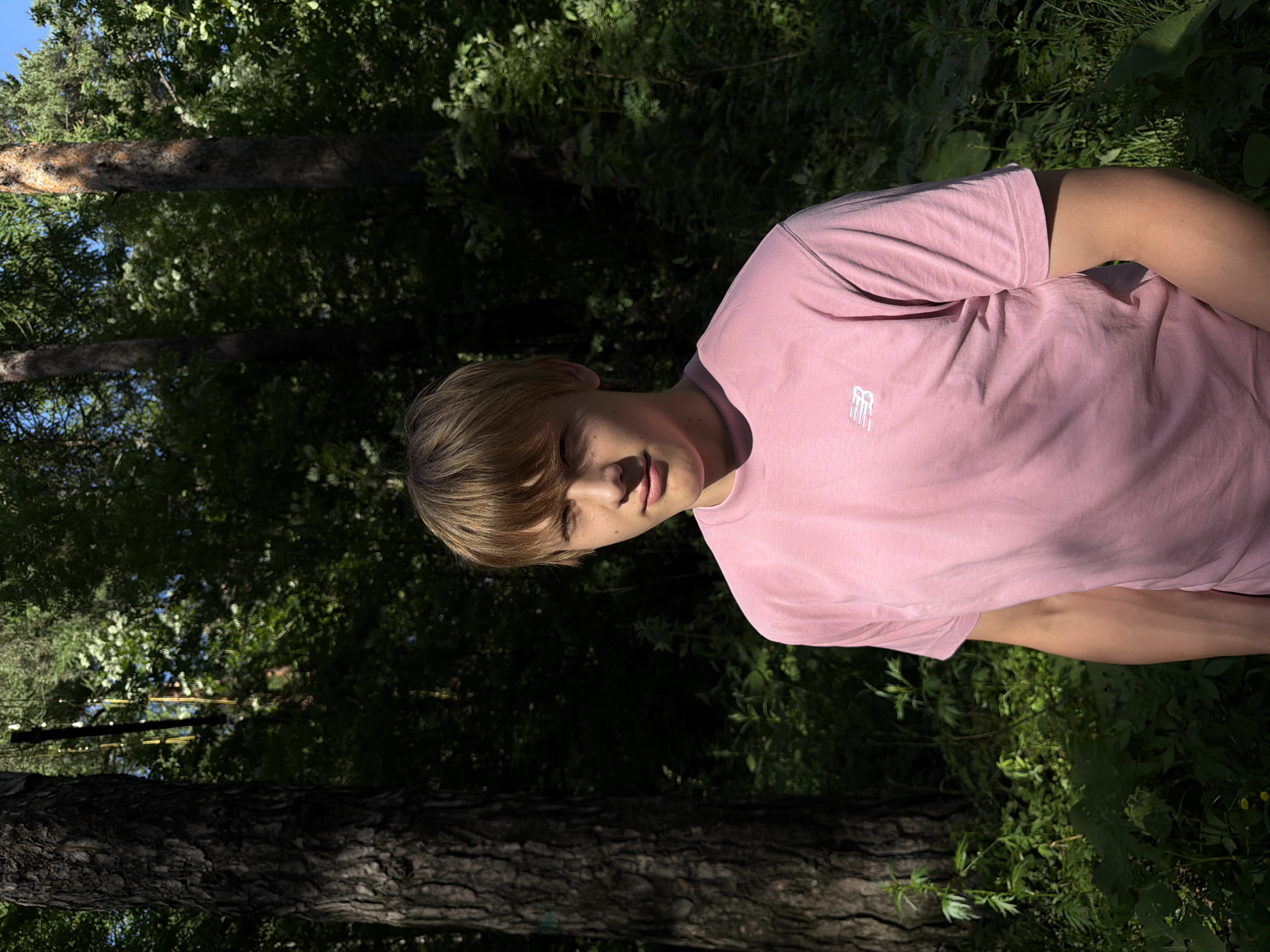 A young person with light brown hair and fair skin is shown from the chest up, their head turned towards the camera. They are wearing a pink t-shirt with a small white logo on the left chest area. The person’s eyes are closed, and their mouth is slightly open, suggesting a relaxed or contemplative pose. Sunlight casts shadows across their face. The background is a dense forest with lush green foliage and tree trunks. The lighting suggests it is daytime, likely late afternoon given the angled sunlight and shadows. The overall impression is a candid or posed portrait set in a natural outdoor environment.