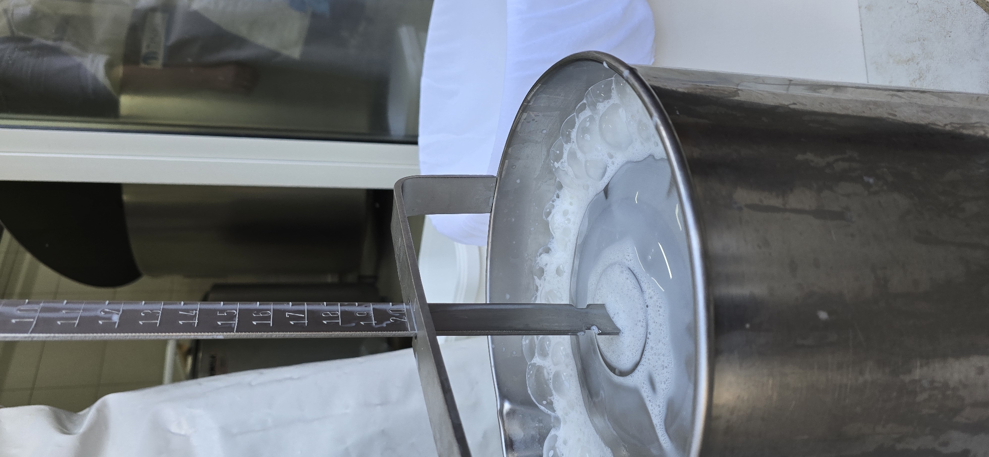 A close-up, slightly tilted photograph shows a stainless steel tank or container partially filled with a milky white liquid with frothy bubbles. A metal ruler or measuring device is positioned across the top of the tank, displaying numbered increments, with the liquid level appearing to correspond to the "19" mark. The setting seems to be indoors, adjacent to a window or similar opening, as hinted by the reflected light and the partial view of an exterior wall. Based on the user’s request, the location is in Blinisht, Albania.