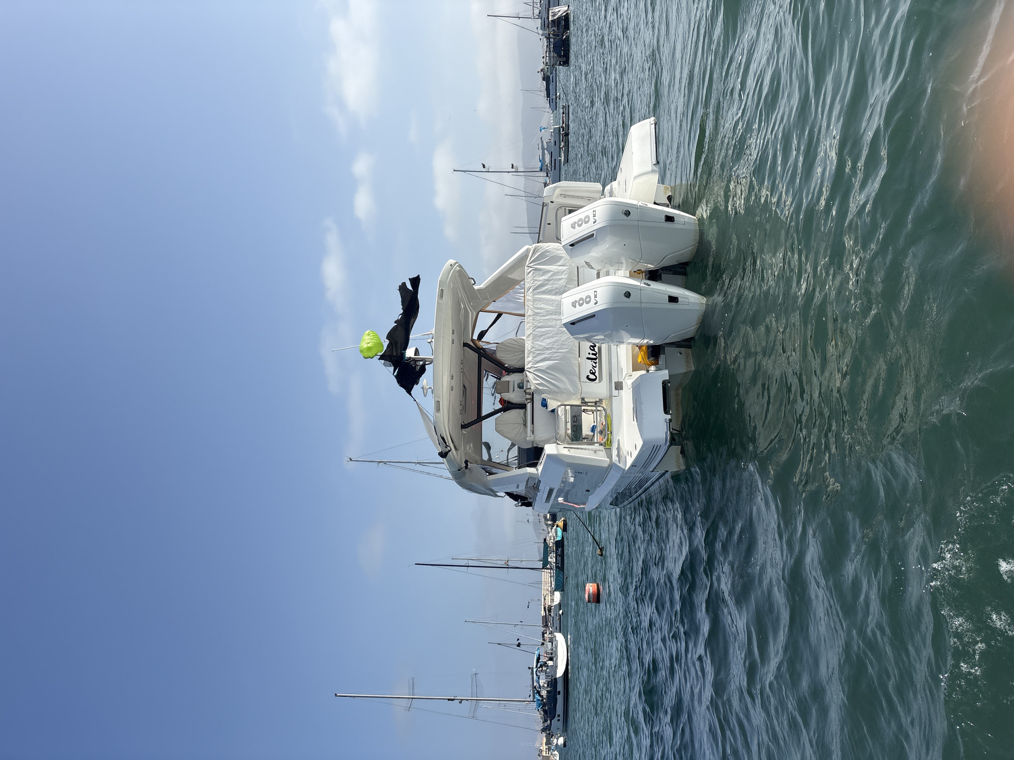 The image depicts a white boat with the name "Cecilia" on its rear, docked in a marina or harbor, likely in La Punta, Peru. The boat has two large outboard motors labeled "400 V10". A green balloon or buoy is visible on a pole extending from the boat. Other boats, including sailboats with masts, are docked in the background. The water is choppy with visible waves. The sky is mostly clear with some scattered clouds.
