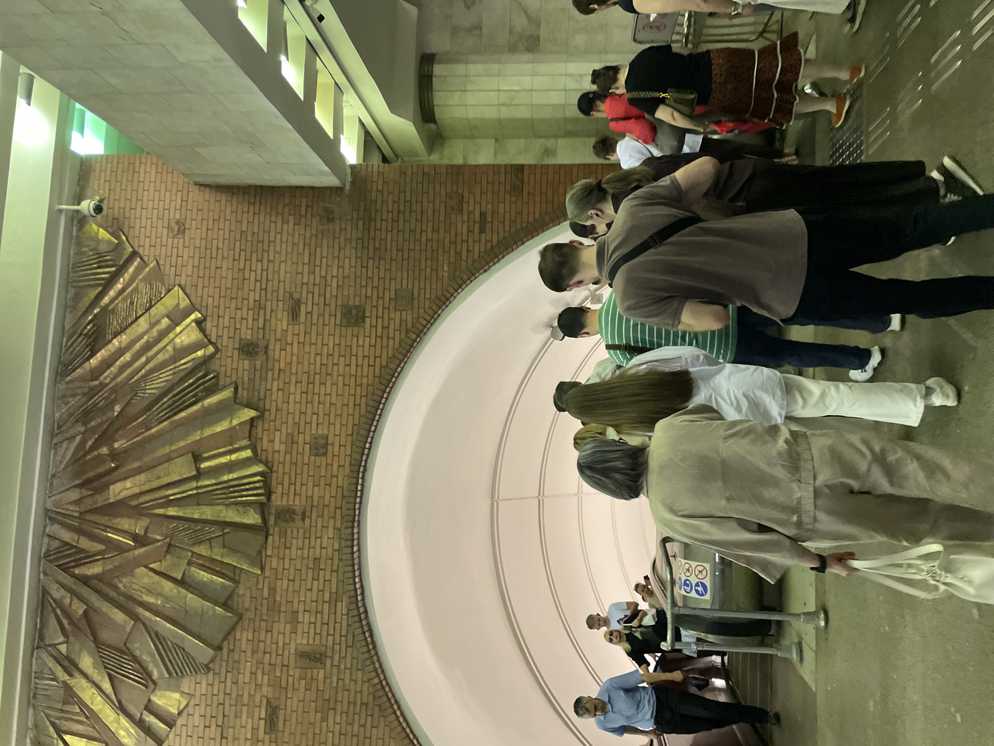 The image captures a scene inside a Moscow metro station in Russia. People are standing, presumably waiting or passing through. The architecture includes brick walls, a large, ornate golden sunburst relief on the wall, and a curved white ceiling. The scene is well-lit and appears to be taken at eye-level.
