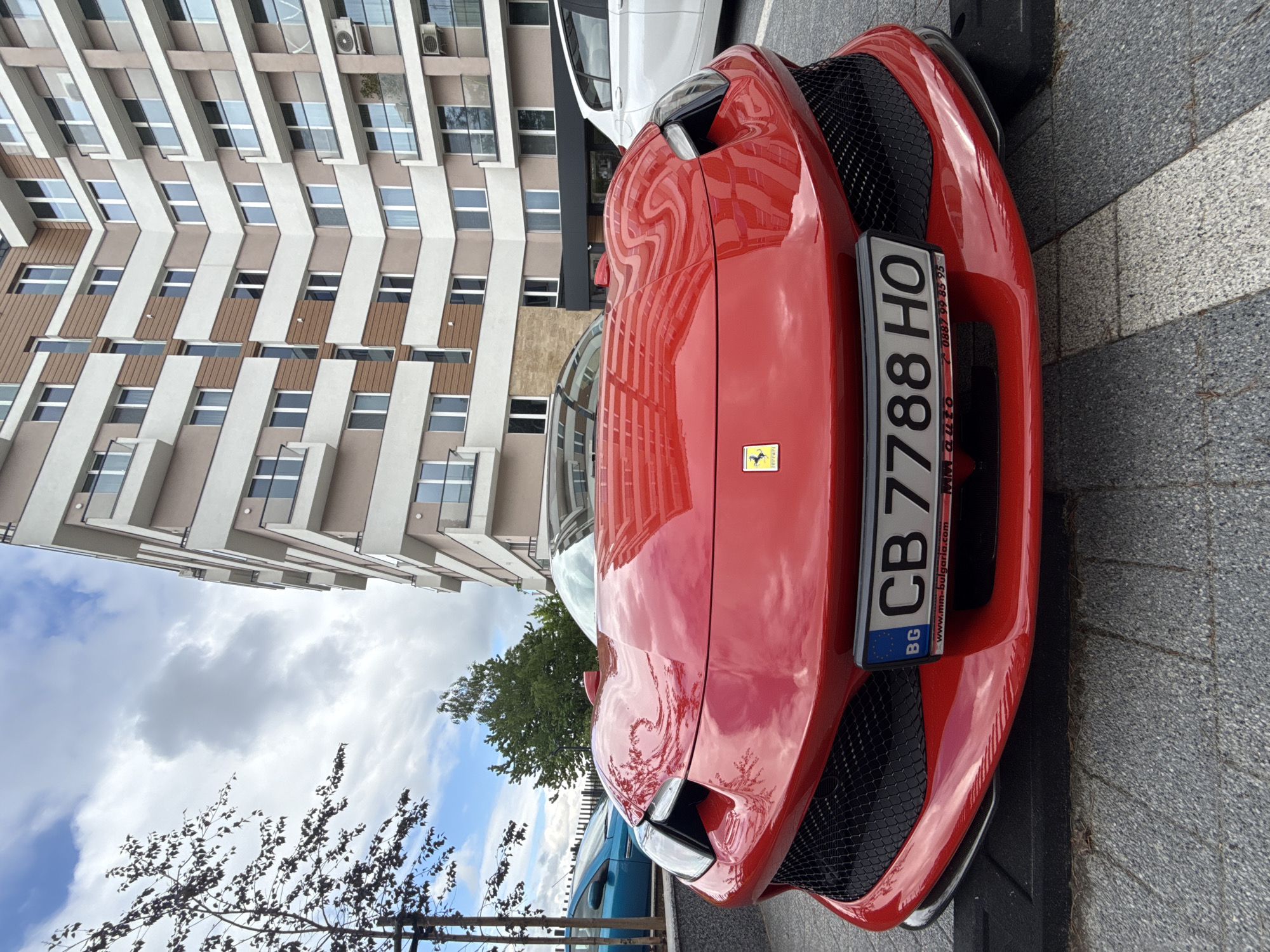 The image shows the front of a red Ferrari parked on a city street. The car has a Bulgarian license plate, "CB 7788 HO". The backdrop features a modern building with a light-colored facade and repeating window patterns. A tree and a patch of cloudy sky are visible in the upper left. The location is likely in Sofia, Bulgaria, as indicated by the license plate and the image's origin. The setting appears to be an urban area. There are no visible people. A white vehicle is parked behind the Ferrari.