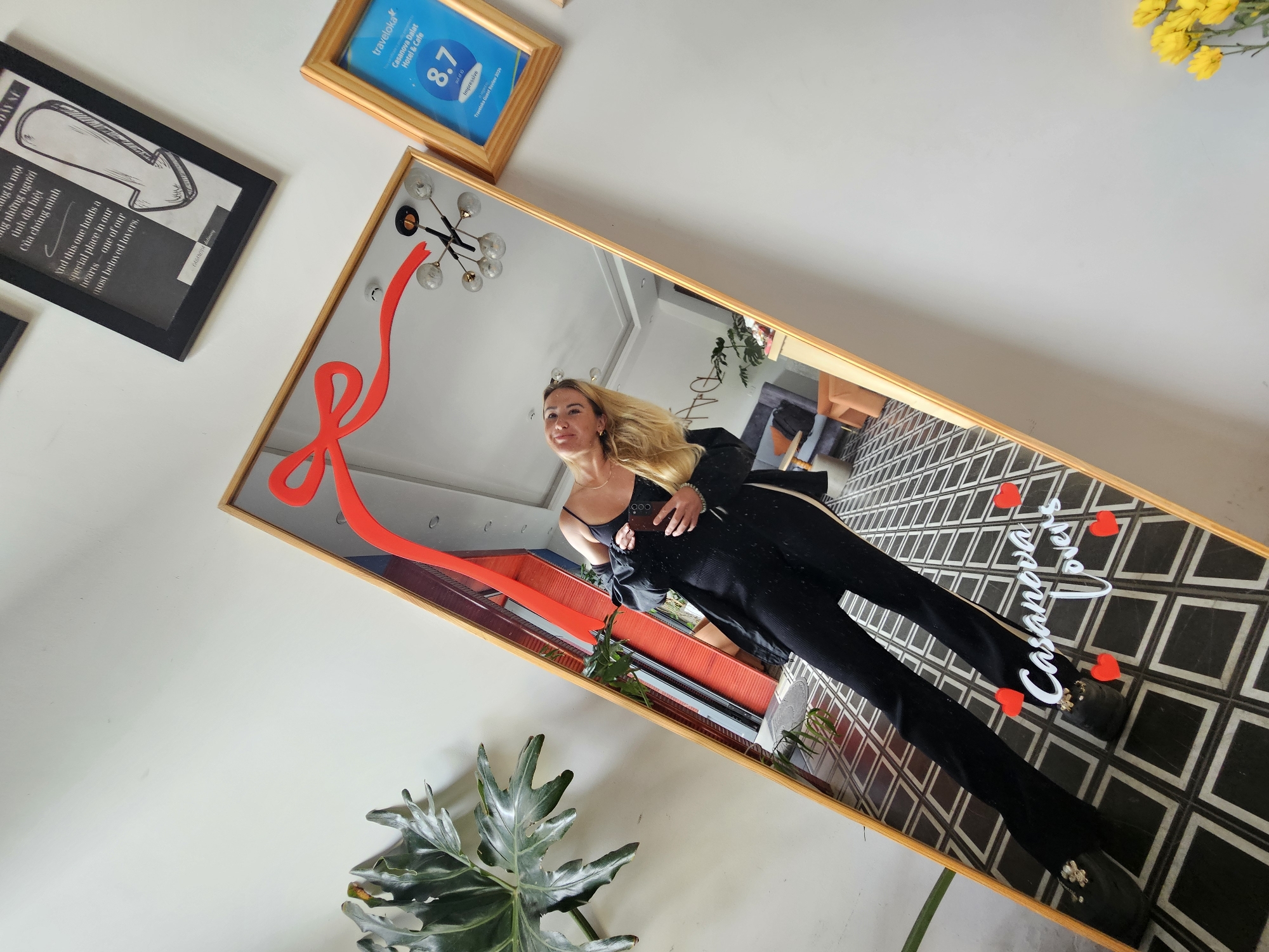 In a hotel lobby in Da Lat, Vietnam, a blonde woman in a black jumpsuit and blazer stands in front of a full-length mirror, taking a selfie. The mirror is decorated with a red ribbon graphic and the words "Casanova Lovers" written in white lettering, surrounded by small red heart stickers. The woman's reflection shows her holding a smartphone. Behind the mirror, a chandelier with multiple bulbs hangs from the ceiling.
On the wall to the right of the mirror, a framed Traveloka review for "Casanova Dalat Hotel & Cafe" displays a score of 8.7, rating it as "Impressive". To the left of the mirror, a large tropical plant with serrated leaves adds a touch of greenery to the scene. Another framed piece of art, with text in Vietnamese and English, hangs above and to the left of the mirror. The floor is tiled with a black and white geometric pattern. The background visible in the mirror shows a modern lounge area with orange seating and a grey sofa. Yellow flowers are visible in the lower right corner of the image, outside the mirror's reflection.