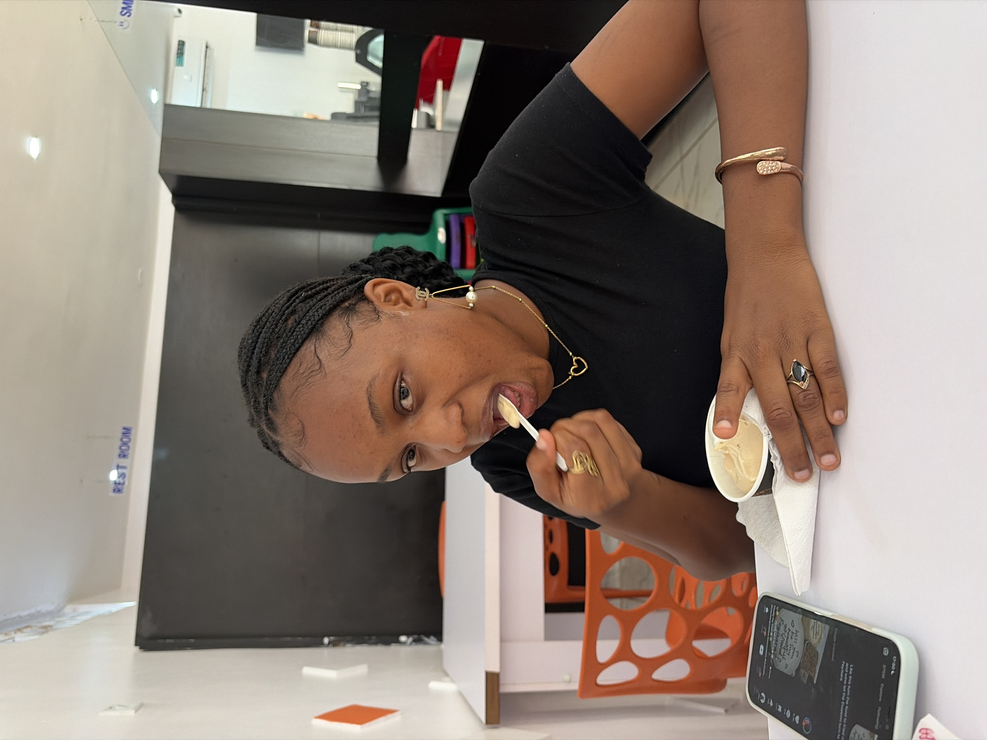 An indoor, eye-level shot, rotated 90 degrees clockwise, captures a woman enjoying a treat. The primary subject, a woman with dark braided hair, is seen mid-bite, holding a small white spoon to her mouth as she eats from a cup of what appears to be ice cream or frozen yogurt. She is looking directly at the camera with an open mouth, suggesting a candid moment.
She is wearing a black short-sleeved top and adorned with jewelry: pearl and gold chain earrings (one with a "CC" logo), a delicate gold chain with a heart pendant, a gold-toned cuff bracelet on her right wrist, and a dark-colored ring on her right ring finger. Her left hand holds the cup, which is wrapped in a white napkin.
The setting appears to be a modern cafe or dessert shop. She is seated at a white table, on which a mobile phone rests in the foreground, displaying a social media interface with the time "17:30" visible. Orange, geometrically designed chairs are visible in the background. Other elements include a dark counter area and a light-colored wall with bright ceiling lights. Visible text includes "SM" in the upper left background on a reflective surface and "REST ROOM" on a distant wall. On the phone screen, snippets of text like "hall you be my Feyi" and "Like this turn the food to..." are discernable.