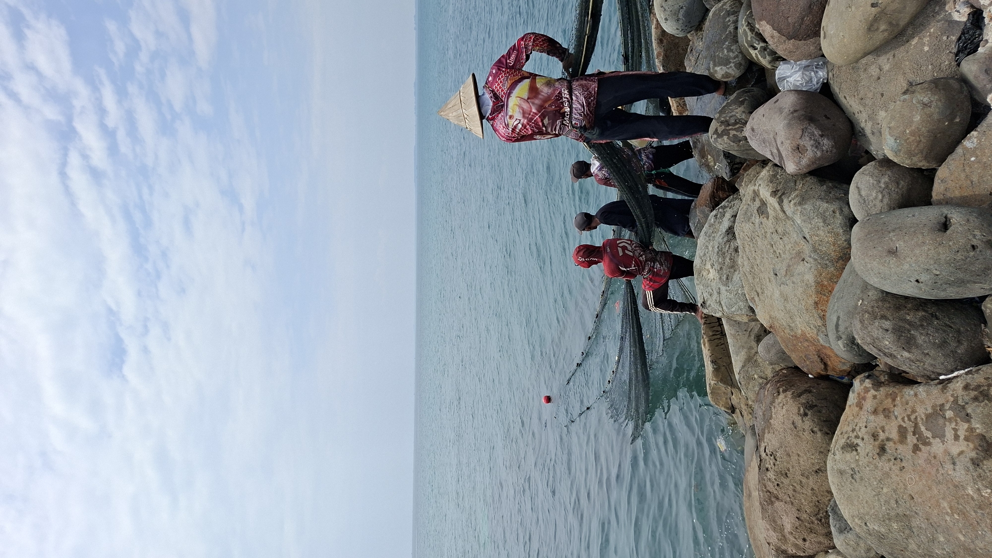This image depicts four individuals engaged in fishing with nets at the rocky shore of a body of water, likely the sea. The scene is set in Lhokseumawe, Indonesia, known for its coastal fishing communities.
In the foreground, large, weathered rocks of varying sizes form the shoreline. The water is a calm, deep blue-green. Several fishing nets are visible, some partially submerged, with a red buoy floating on the surface.
The four individuals are positioned near the water's edge, actively handling the nets. One person, facing away from the camera, wears a conical straw hat and a brightly colored shirt featuring a fish motif and text. Another individual, also facing away, is dressed in a red hooded jacket with black and white stripes on the sleeves and pants, leaning forward as if pulling a net. A third person, wearing a dark shirt and a grey baseball cap, stands beside them. A fourth person, partially obscured, is also involved in the fishing activity.
The sky above is a pale blue, dotted with scattered white clouds, suggesting a bright, possibly overcast day. The overall impression is one of a typical fishing scene in a coastal Indonesian city, emphasizing the communal effort involved in the activity.