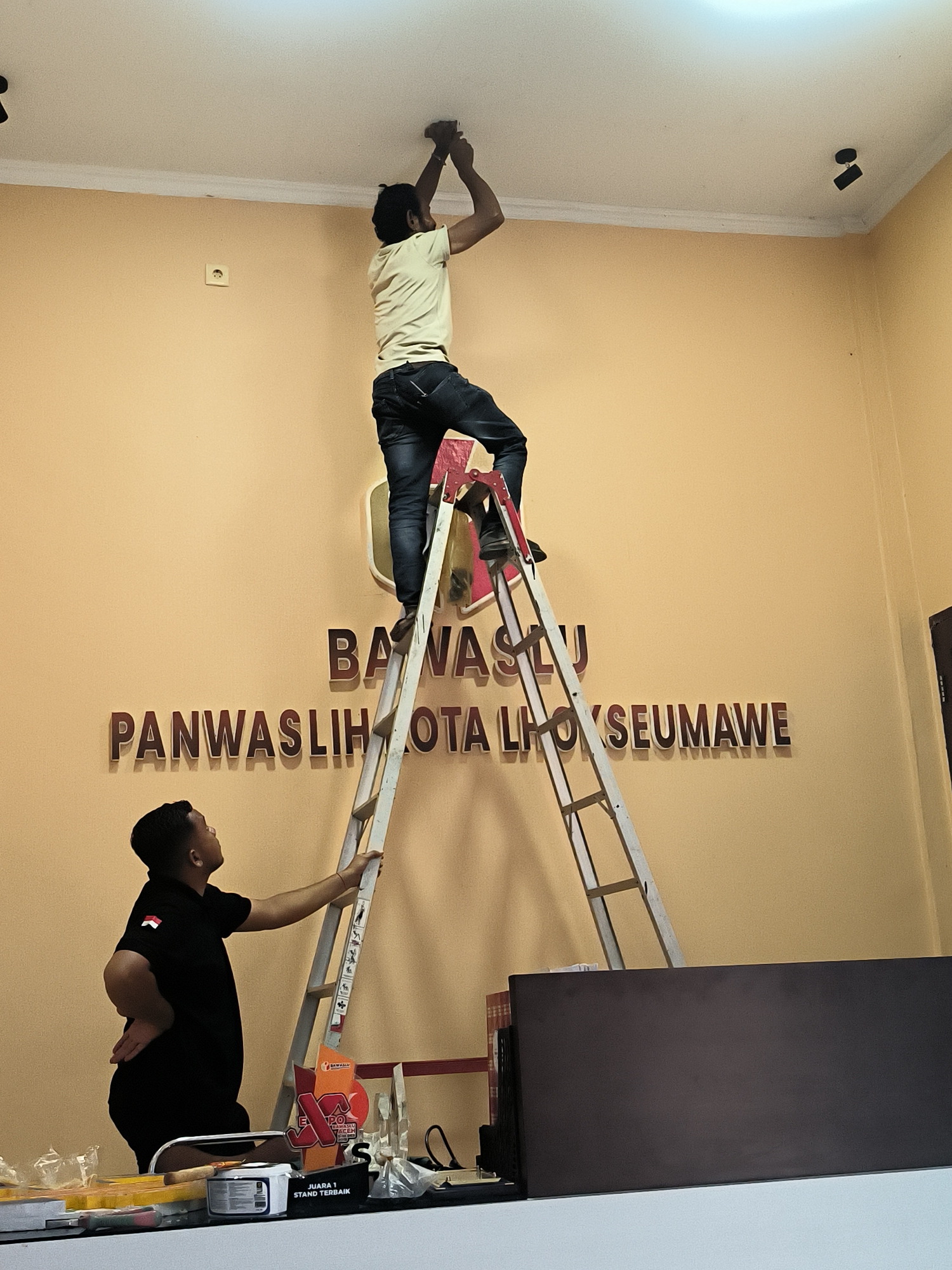 Two men are working on a wall in a room with light orange walls. The man on the right is standing on a tall, A-frame ladder, reaching up to the ceiling to work on a light fixture. He is wearing a light yellow t-shirt and dark jeans. The man on the left is standing on a shorter ladder, looking up at the man on the taller ladder. He is wearing a black t-shirt with an Indonesian flag patch on the left sleeve. Behind the men, a large, 3D sign on the wall reads "BAWASLU PANWASHLIHOTA LHOKSEUMAWE." The room appears to be an office or public space, with some office furniture and materials visible in the foreground.
