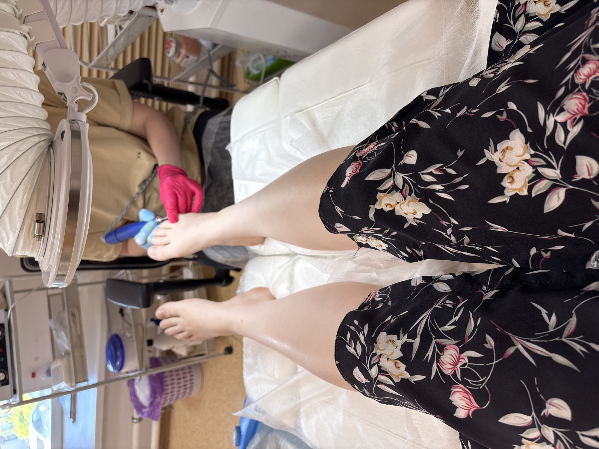 A person wearing a black floral dress lies on a white treatment table. Their bare legs extend towards the center of the frame. A medical professional, wearing pink gloves and a tan shirt, is performing a procedure on the patient's left foot with a blue instrument. Medical equipment, including a white ventilation system and a metal cart, are visible. The location is possibly Chaikovsky, Russia, based on prior instruction.