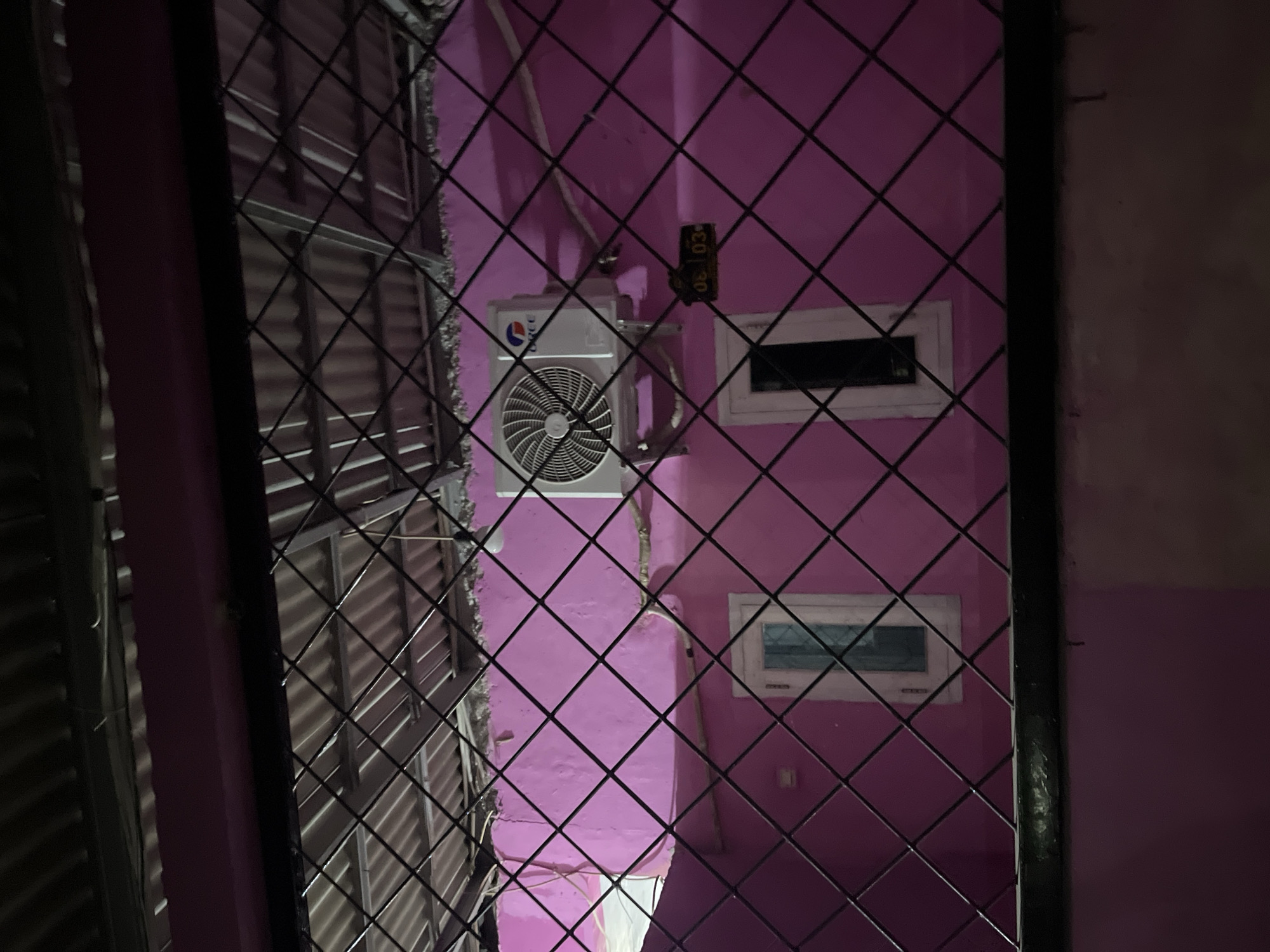 The image depicts a pink-colored exterior wall of a building, viewed through a black metal grate. An air conditioning unit is mounted on the upper portion of the wall, to the right of center. Below the air conditioner, a small digital display shows "06 03". Two rectangular windows, one with a visible pane and the other dark, are present on the wall. The scene is dimly lit, suggesting it is nighttime. The image is a view from an elevated position looking down onto the wall. The location is identified as Laban, Indonesia.