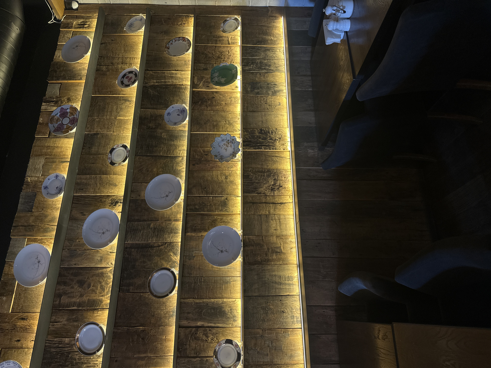 The image depicts a decorated wall and adjacent seating area, possibly inside a restaurant. The wall is constructed of horizontal wooden planks, illuminated by vertical light strips. Decorative plates of varying designs are mounted on the wall. To the right of the wall is a dimly lit seating area with padded chairs or benches and a table. The location is likely Izhevsk, Russia. There are no people visible in the image.