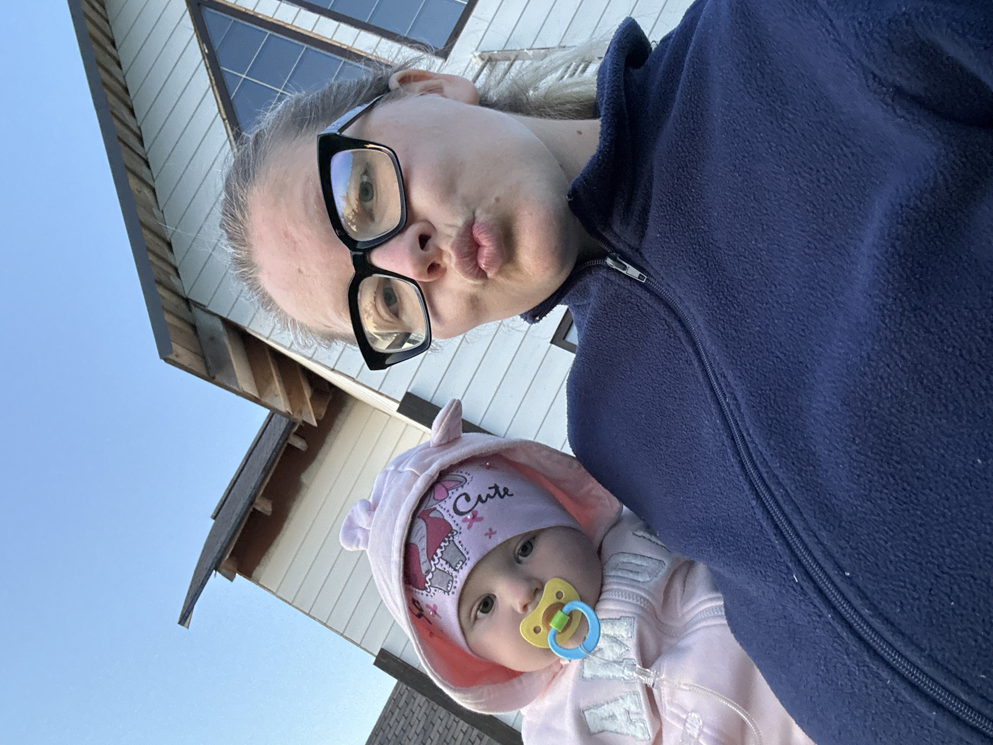 A medium shot captured from a low angle shows a woman and a baby posing for a selfie outdoors. The woman, wearing a dark blue fleece jacket and black-rimmed glasses, puckers her lips. She is holding a baby, who is wearing a pink hooded jacket and a pink beanie with an elephant design and the word "Cute" on it. The baby has a yellow pacifier in its mouth. In the background, a light-colored house with a dark roof and a large triangular window is visible. The sky is clear and blue. The image appears to have been taken in Abushkovo, Russia.