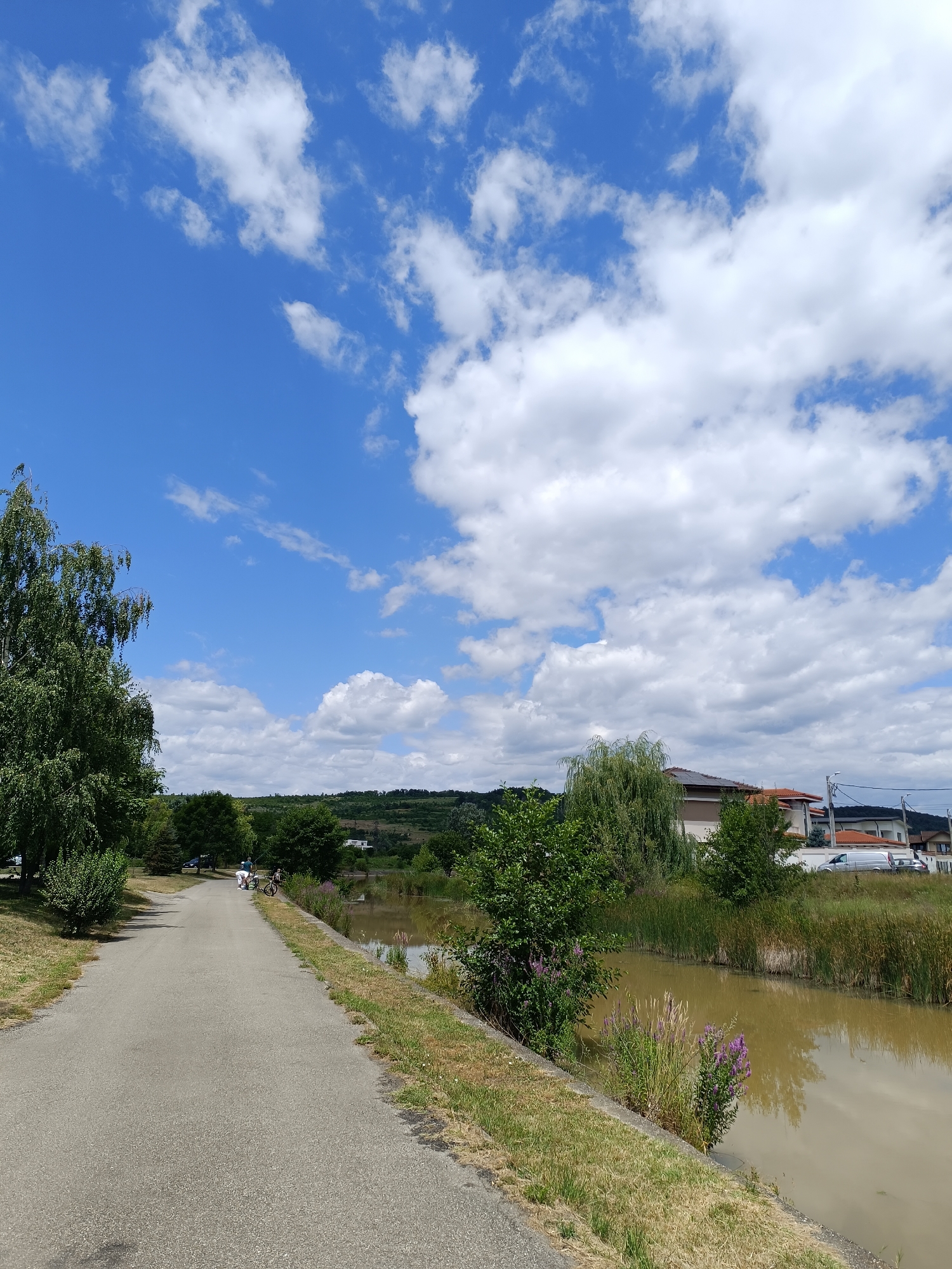 A bright, sunny day with blue skies and scattered white clouds is depicted. A paved path runs alongside a murky, brown canal. Lush green trees and vegetation line the left side of the path and the canal. In the background, there are houses, some trees, and rolling hills. A few people are visible on the path, one person on a bicycle and two others standing near it. A car is parked on the left side of the path. The canal reflects the sky and surrounding greenery. The scene suggests a peaceful, outdoor recreational area, likely within or near the city of Târgu Jiu, Romania, given the architectural style of the houses and the general landscape.