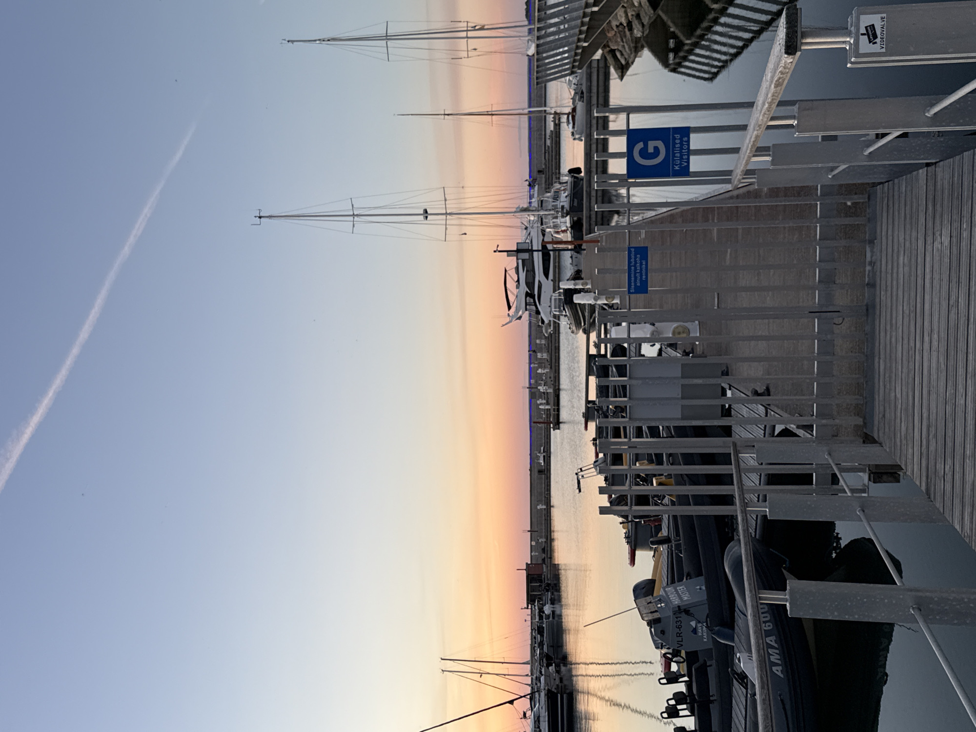 The image depicts a marina in Tallinn, Estonia, at sunset. The sky is a gradient of pale blue to warm orange, with a faint contrail from an aircraft visible. Several sailboats and motor yachts are docked in the calm water, their reflections visible on the surface. A wooden pier with metal railings leads into the marina. Signs on the pier indicate restricted access, with one in Estonian and English stating "Entrance is permitted only to the berth holder" and another with a "G" symbol and "Külalised Visitors" written below it. There are no people visible in the scene. The overall atmosphere is serene and quiet, characteristic of a marina at the end of the day.