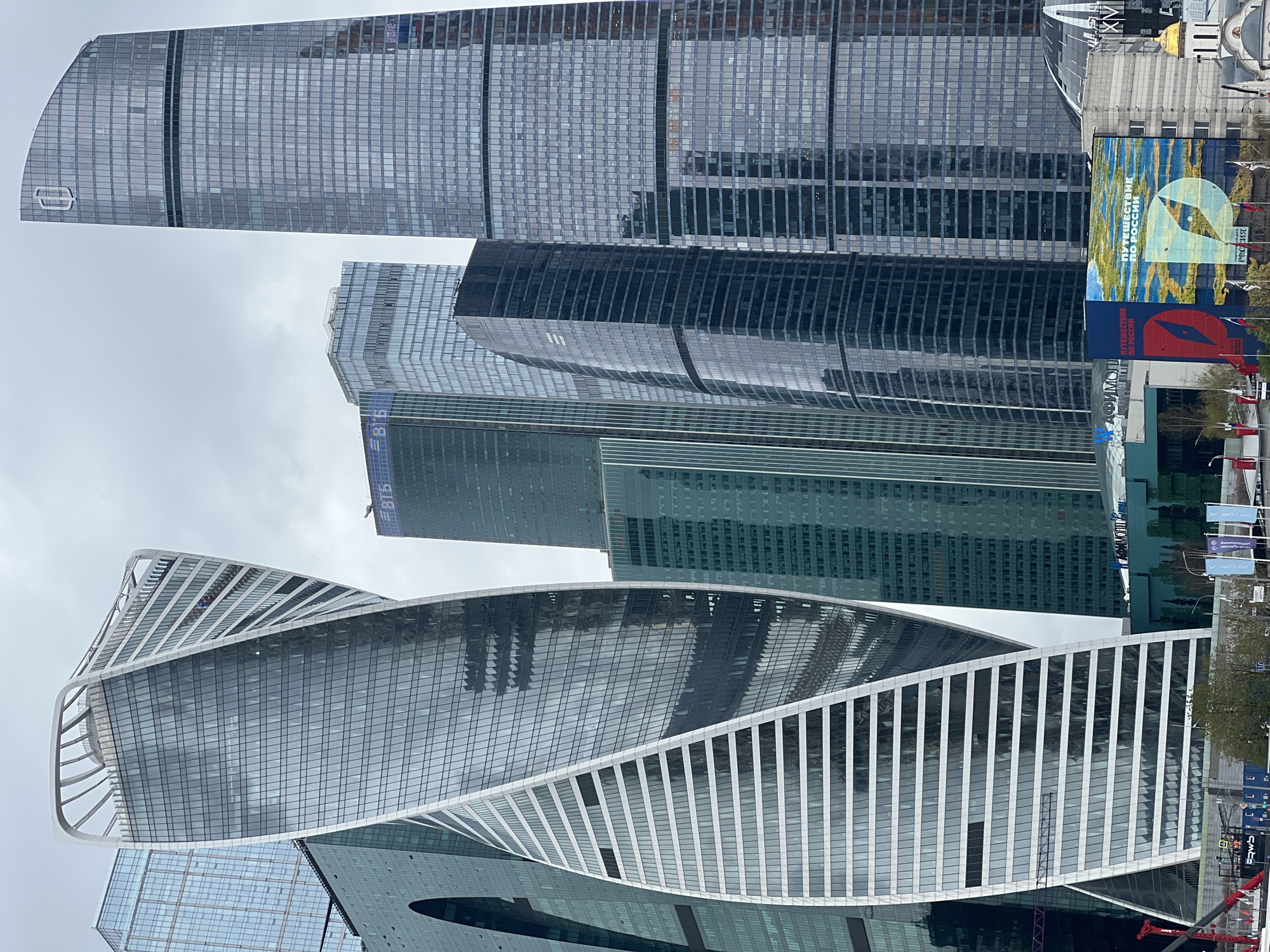 The image shows a cluster of modern skyscrapers in Moscow, Russia, specifically within the Moscow International Business Center (MIBC), also known as Moscow City. The dominant building in the foreground is the Evolution Tower, distinguished by its spiraling shape and white exoskeleton. Behind it are other skyscrapers of varying heights and designs, with glass facades reflecting the overcast sky. To the right of the buildings is a street, with banners and signs visible. A large sign shows a compass with the Russian words "Путешествие по России" which translates to "Travel Across Russia". There are also some trees and smaller buildings to the right of the skyscrapers. The overall impression is one of a dense urban area characterized by modern architecture.