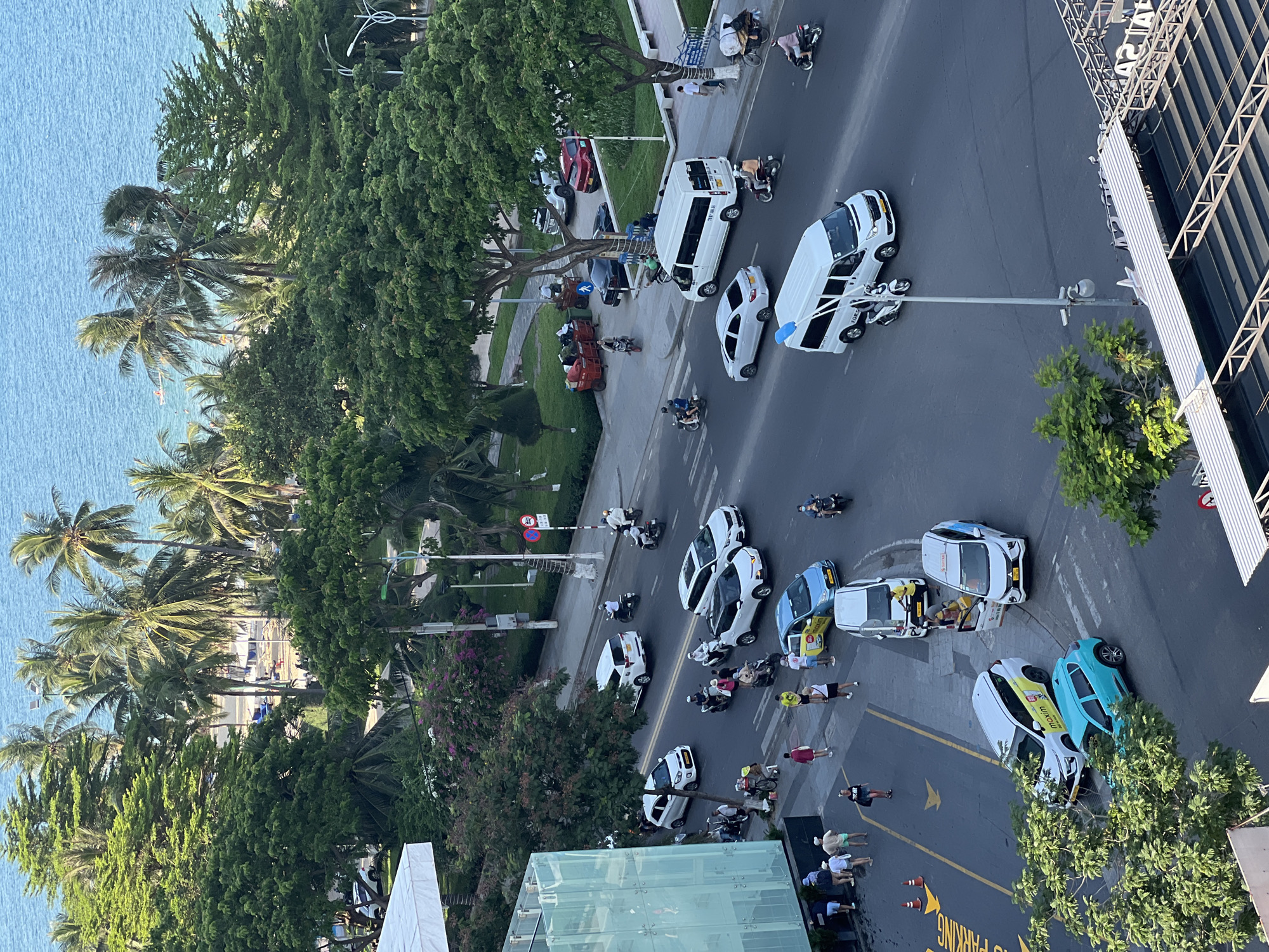 An aerial shot captures a street scene in Nha Trang, Vietnam, with a body of water and palm trees visible in the background. Cars, motorbikes, and vans navigate a multi-lane road, while pedestrians walk along the sidewalk. A few people are congregated near a food stall. The scene appears to be a typical day with moderate traffic and activity.