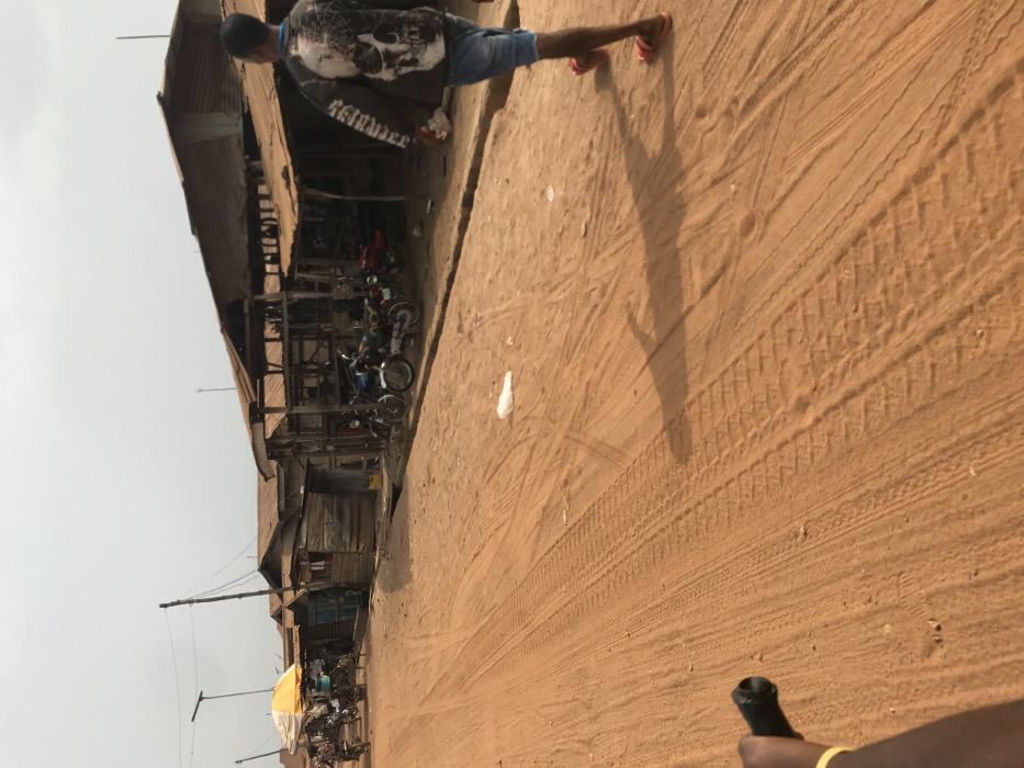 The scene depicts an unpaved, reddish-brown earth road in Ogbese, Nigeria, under an overcast or hazy sky. A row of weathered, single-story structures with corrugated metal roofs and wooden walls lines the right side of the road. Several of these structures appear to be open-fronted stalls, with multiple motorcycles parked in front of them. The road surface exhibits numerous tire tracks and foot impressions. A white plastic bag lies on the ground in the mid-distance.
A male individual is observed from behind, walking away from the camera on the right side of the road. He wears a dark long-sleeved shirt featuring a large white skull graphic on the back, denim shorts, and red flip-flops. His shadow is cast prominently on the ground to his left. In the far left distance, several indistinct figures are gathered near a yellow market umbrella, with additional vehicles parked nearby. A human hand, wearing a yellow bracelet and gripping a black handlebar, is visible in the lower-left foreground, indicating the perspective of a person on a bicycle or motorcycle. Utility poles with overhead wires run along the left side of the road.