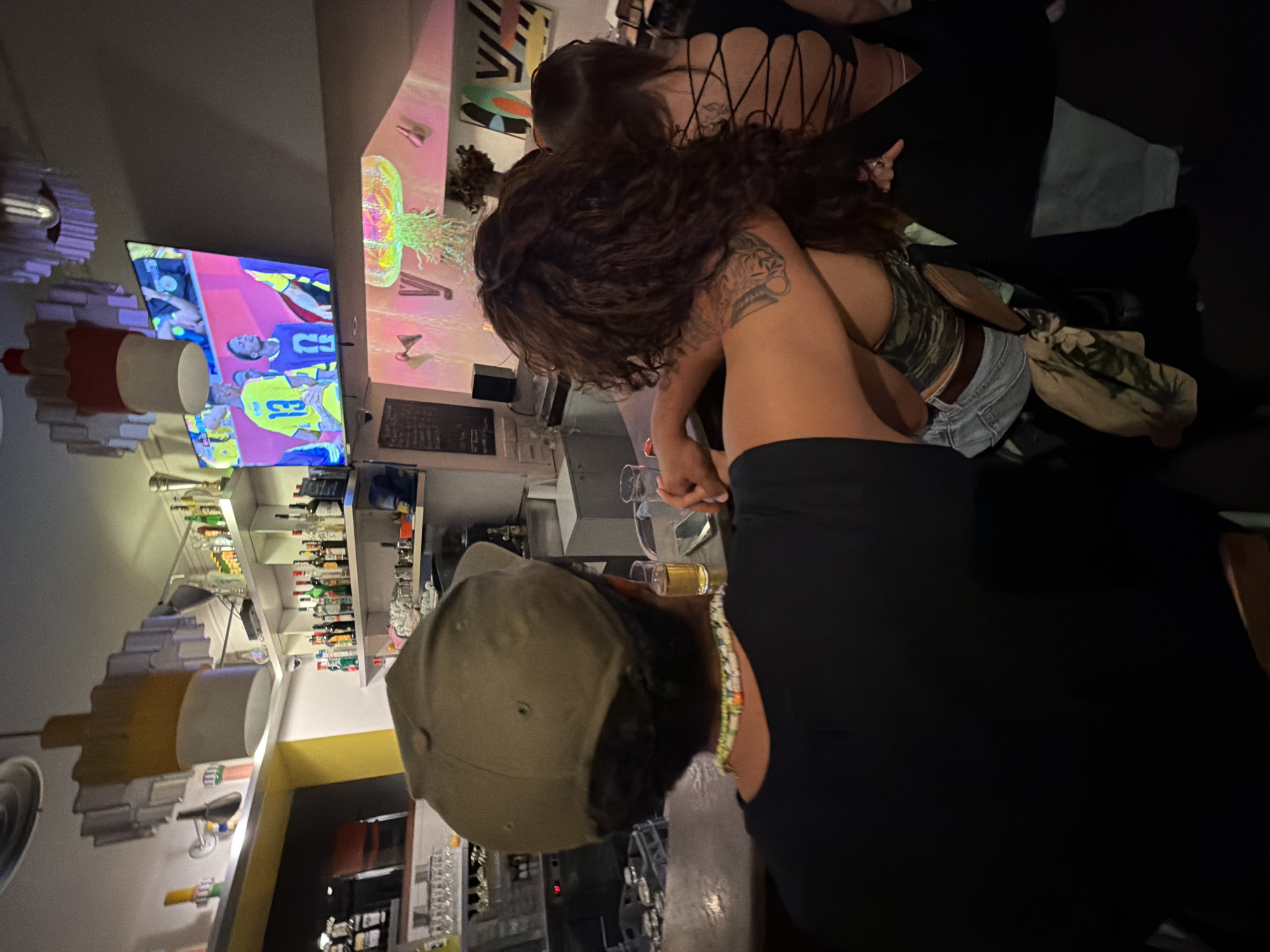 The image shows three individuals at a bar, with a television screen displaying a sports game in the background. A person wearing a green baseball cap is seated at the bar, facing away from the camera, with their arm around another individual with long, curly brown hair. This second individual is wearing a camouflage-patterned top and denim shorts, and their arm is resting on the first person's shoulder. To the right, another person with dark hair is visible, wearing a black top with a criss-cross back design. The bar area appears to be indoors, with shelves of bottles and glasses visible behind the individuals. The overall lighting is dim, creating a relaxed atmosphere. No specific landmarks or elements clearly identify the location as Lyon, France.