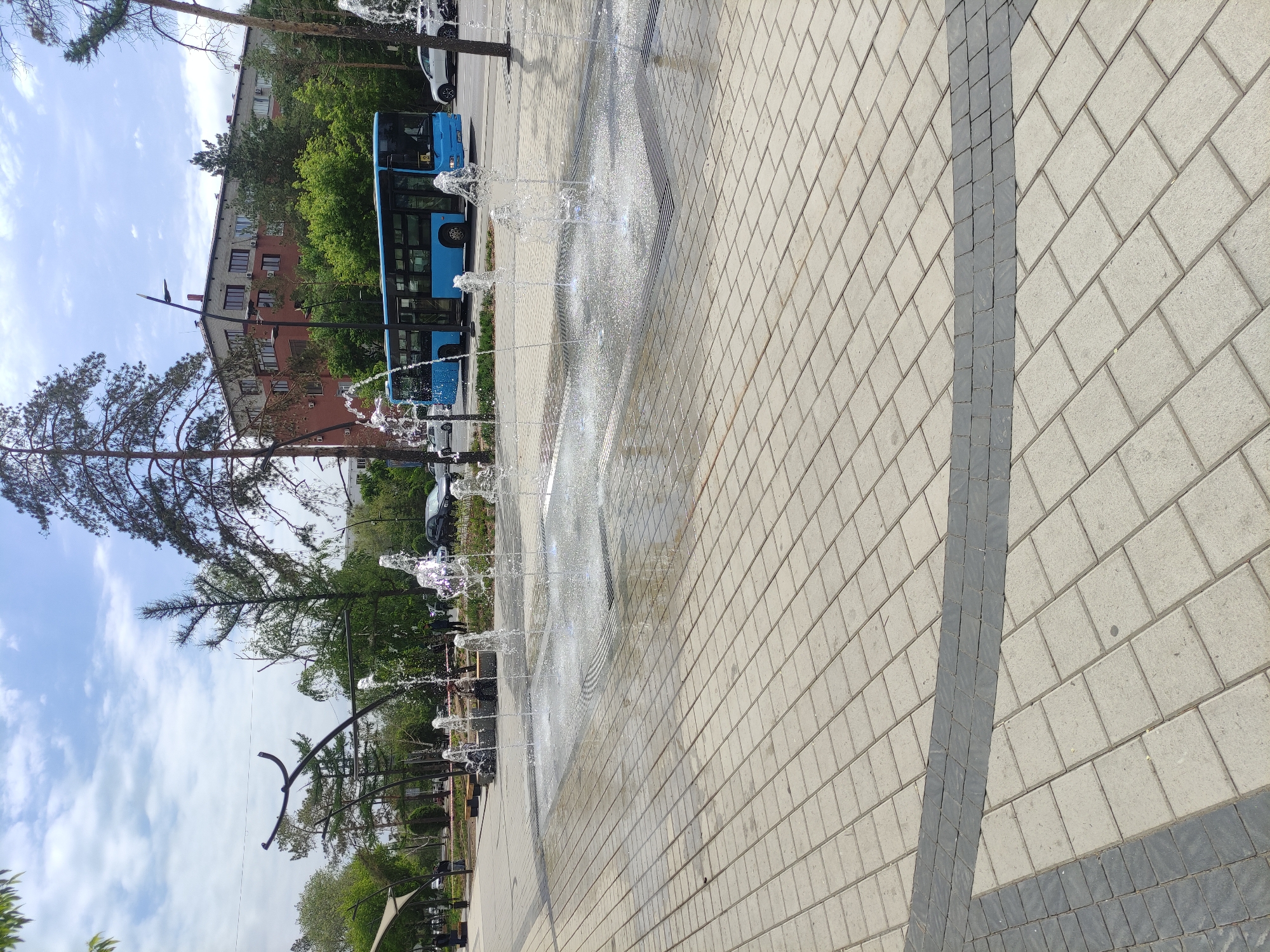 The image captures a scene in Pavlodar, Kazakhstan. A paved plaza, constructed from interlocking bricks in a herringbone pattern, dominates the foreground. A row of water fountains is positioned alongside the plaza.
In the background, a blue bus is visible, indicating urban transit. A residential building stands further behind the bus. Trees are interspersed throughout the background, adding greenery. The sky is partially cloudy.
No people are visible in the image. There are no explicit actions or interactions. The overall environment appears to be an urban public space.