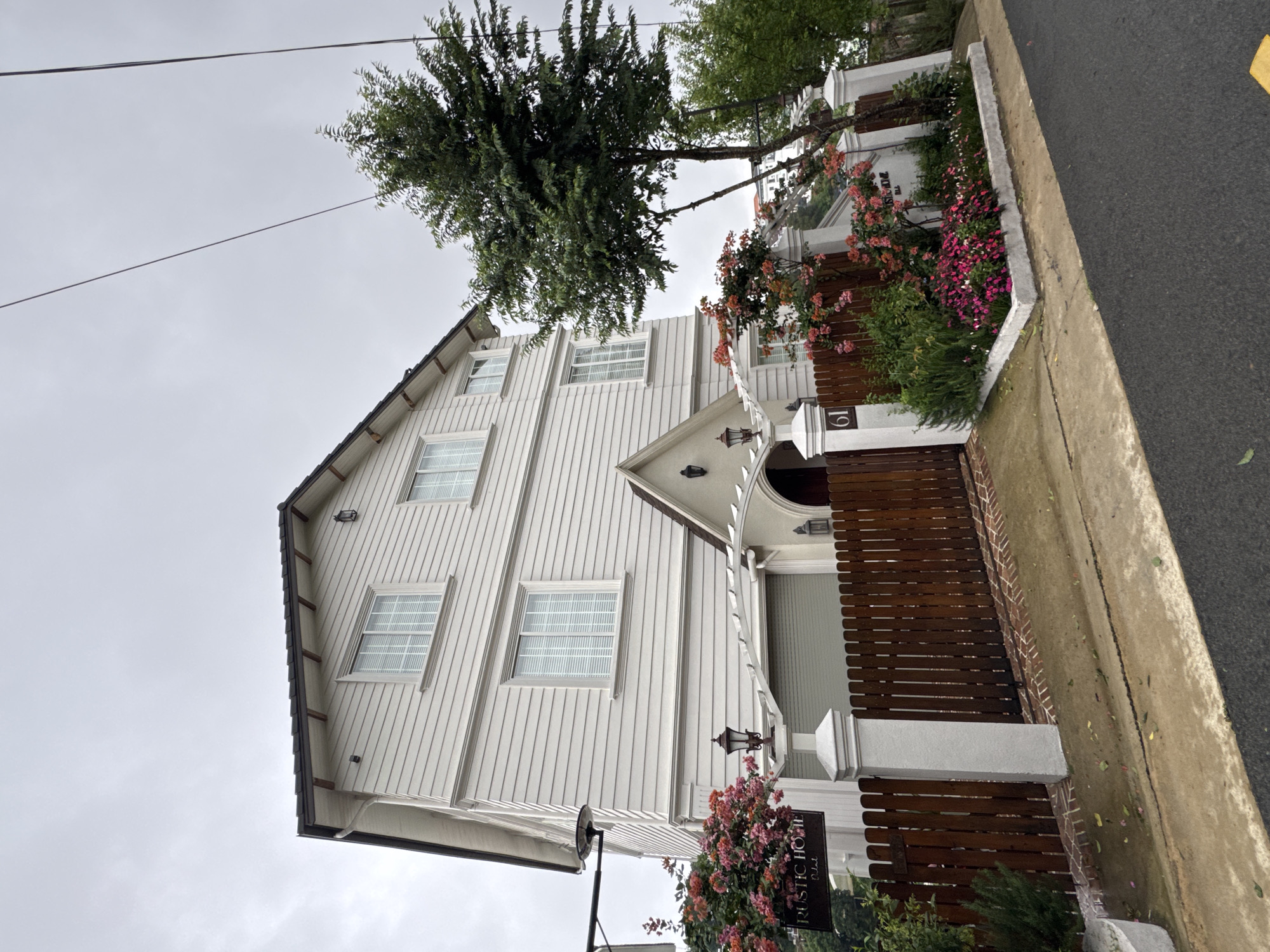 An upright-oriented shot captures the exterior of a charming, multi-story white house with a dark roof under an overcast sky. The house features vertical wooden siding and multiple windows with white frames, each fitted with horizontal blinds.
In the foreground, a well-maintained garden bursts with colorful pink and reddish flowers and lush green foliage, bordering a dark wooden fence. A tall, leafy green tree stands prominently to the left of the house, its branches extending upwards and to the right, partially obscuring the upper sections of the building. Utility lines stretch across the dull grey sky above.
A paved sidewalk and a dark asphalt road are visible on the far right, suggesting the property is situated along a street. Notable details include the house number "61" displayed on a white pillar near the entrance gate. A black sign hangs from a white post within the garden, clearly displaying the text "RUSTIC HOME Hotel" in white lettering, indicating the building's function. An ornate black lantern is mounted above the arched entrance of the house. The overall impression is of a quaint and inviting property, possibly a guesthouse or small hotel, located in Lam Vien - Da Lat, Vietnam.