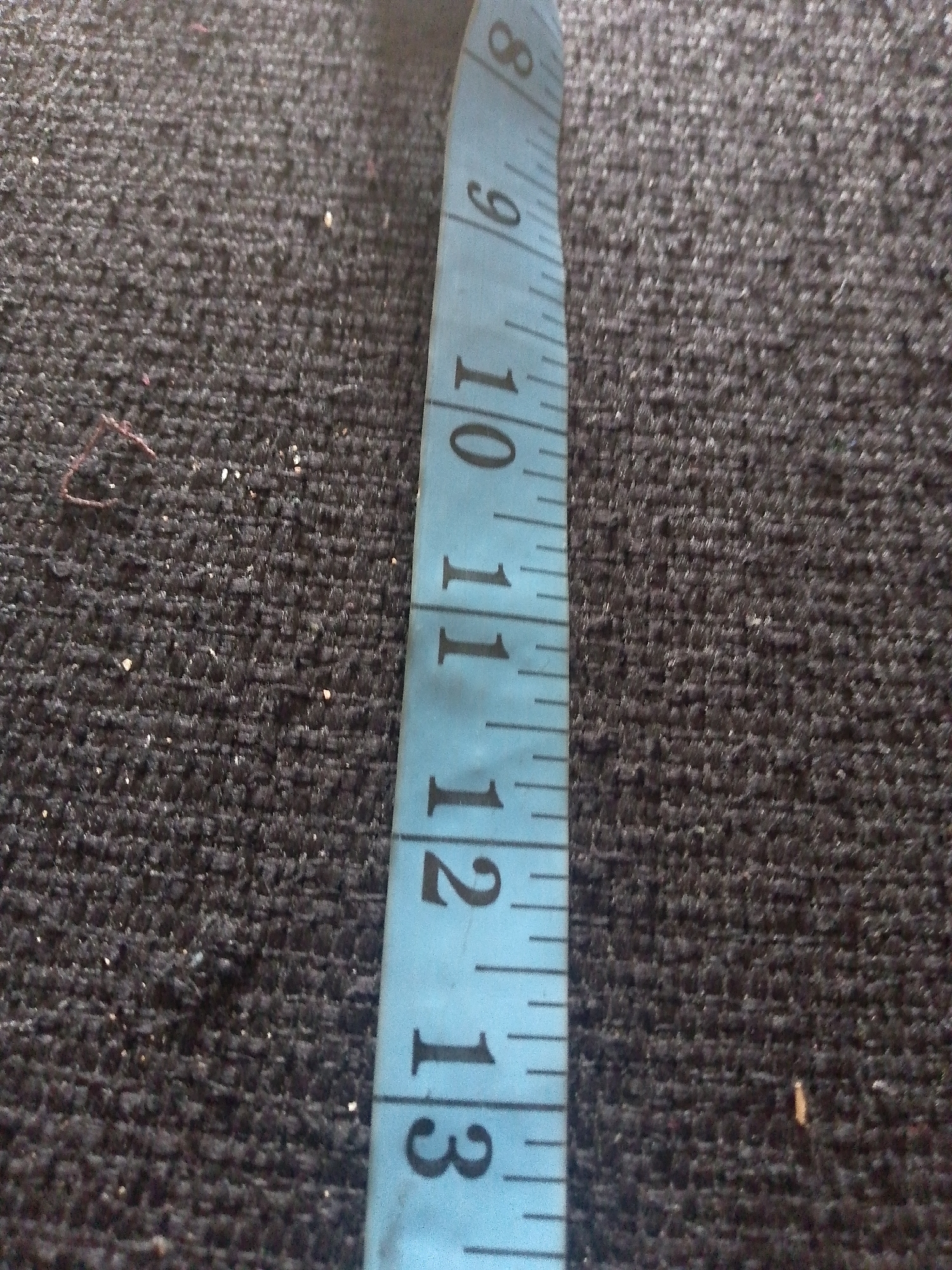A close-up shot shows a blue measuring tape with black markings on a dark, textured surface. The tape is oriented vertically and diagonally from the top right to the bottom left of the frame. Markings visible include numbers 8, 9, 10, 11, 12, and 13, along with smaller tick marks indicating inches or centimeters. The background surface appears to be a carpet or a similar fabric, with a rough, woven texture and some small white and reddish specks scattered across it. The lighting is uneven, casting subtle shadows and highlighting the texture of the surface. There are no people or animals visible in the image, nor any context to suggest a specific location within Jauro Yinu, Nigeria, beyond the provided prompt.