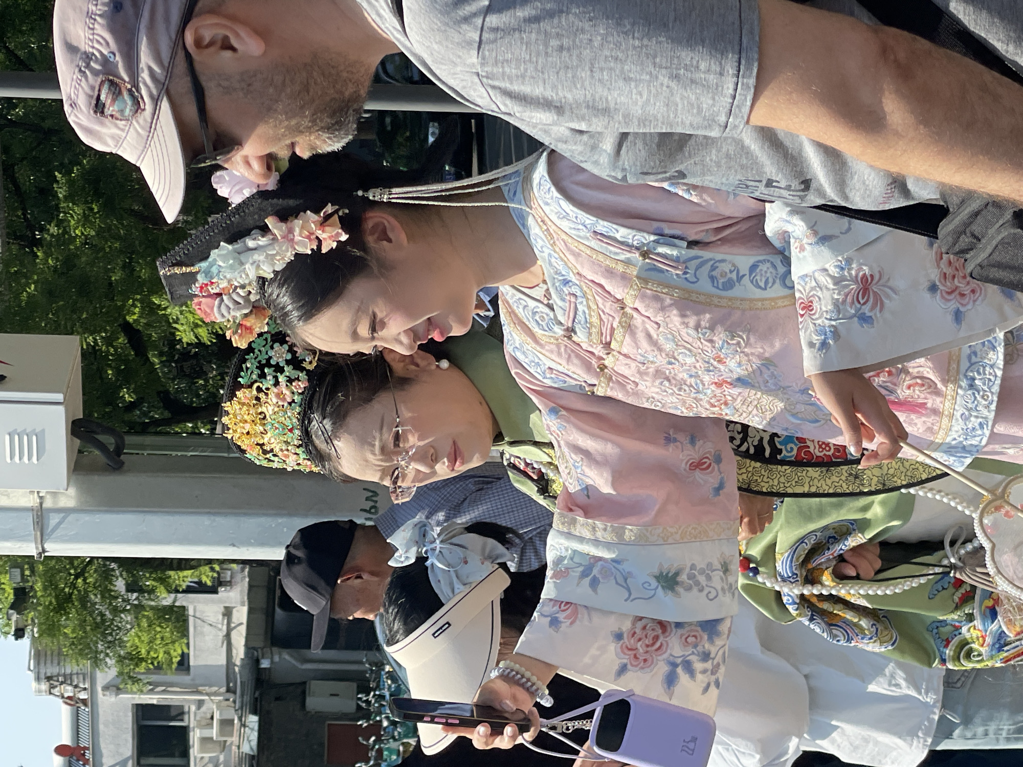 The image depicts two women and two men outside, likely in the Dongcheng District, China. The women are wearing traditional Chinese clothing with ornate headpieces. One woman is in a light pink dress, the other is wearing a green dress. The men are dressed in casual western clothing. The woman in green is holding a phone. A partial view of buildings and trees can be seen in the background.