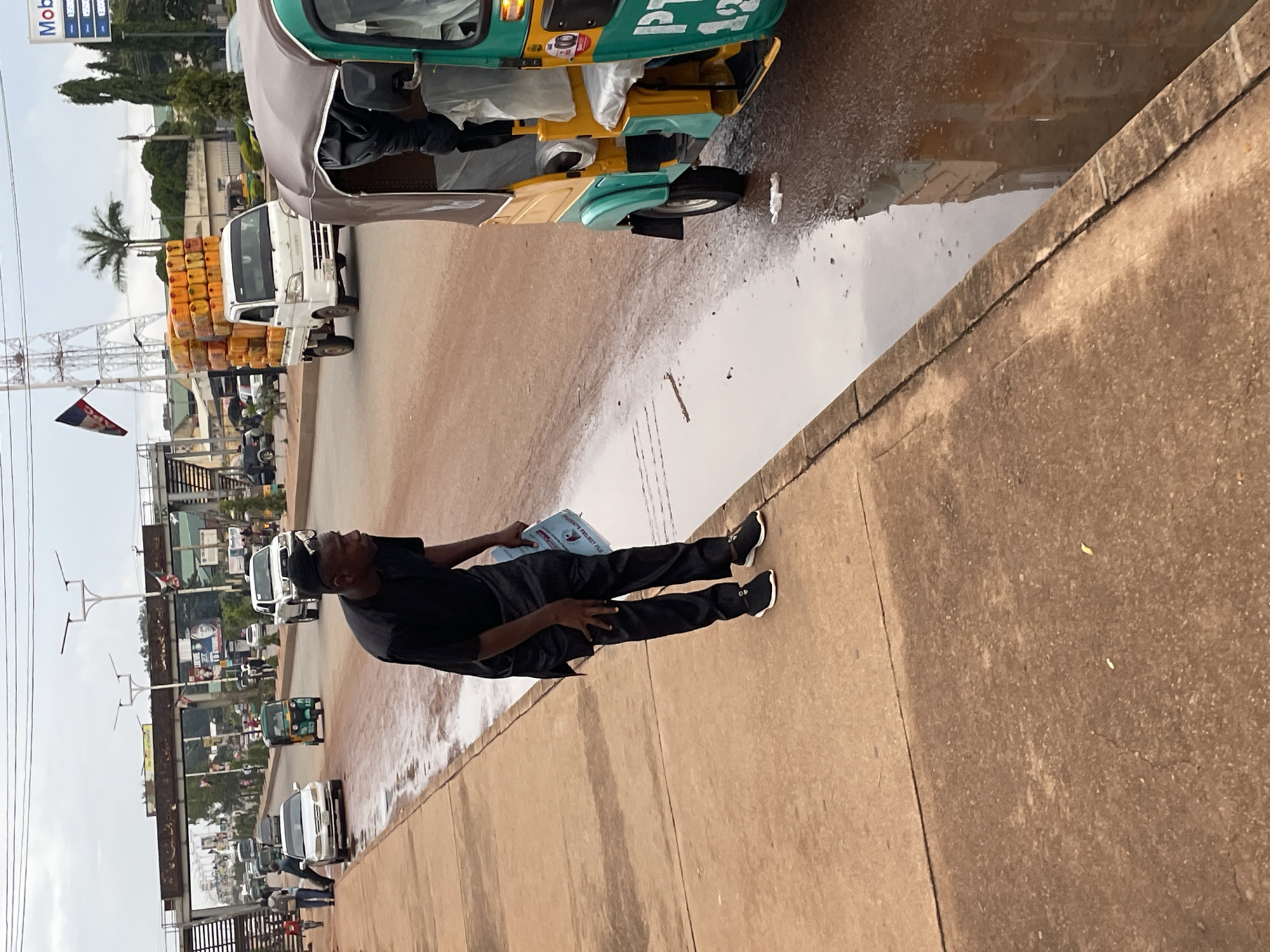 A man in a black traditional outfit stands on the side of a wet, reddish-brown road in Jos, Nigeria, holding a blue document. He wears black sneakers and has a distinctive haircut. In the foreground, a yellow and green auto-rickshaw, commonly known as a "Keke Napep," is parked. The road is partially covered in standing water, indicating recent rain.
In the background, other vehicles are visible on the road, including a white truck heavily loaded with yellow jerrycans, a silver SUV, a white sedan, and a green auto-rickshaw. An overpass with signs and flags crosses the road. Buildings and trees line the street, and a cell tower stands tall in the distance. The sky is overcast, suggesting a cloudy day. The scene appears to be a busy street with ongoing traffic and pedestrian activity.