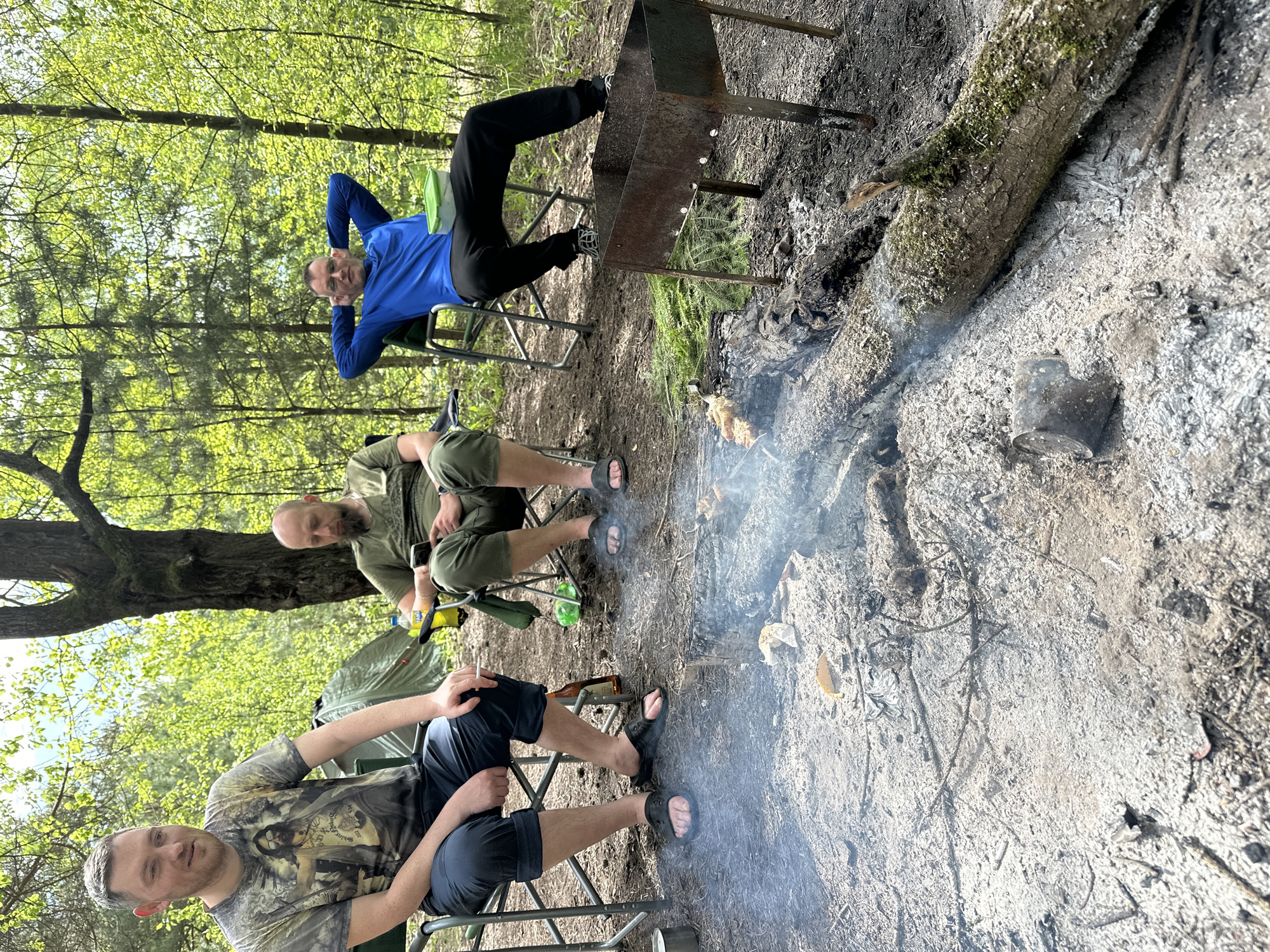 The image shows three men sitting around a makeshift grill in a forest, possibly in the area of Юзуфоўскі сельскі Савет, Belarus. One man, wearing a blue shirt, is reclining in a chair with his hands behind his head. Another man, dressed in an olive-green outfit, is seated in a chair and looking down. The third man, wearing a printed t-shirt, is sitting in the foreground looking toward the camera. A grill with skewers above is visible next to the fire. There is ash covering the ground, indicating a recent fire. A log lies nearby. There is a tent in the background. The lighting is bright, suggesting daytime.