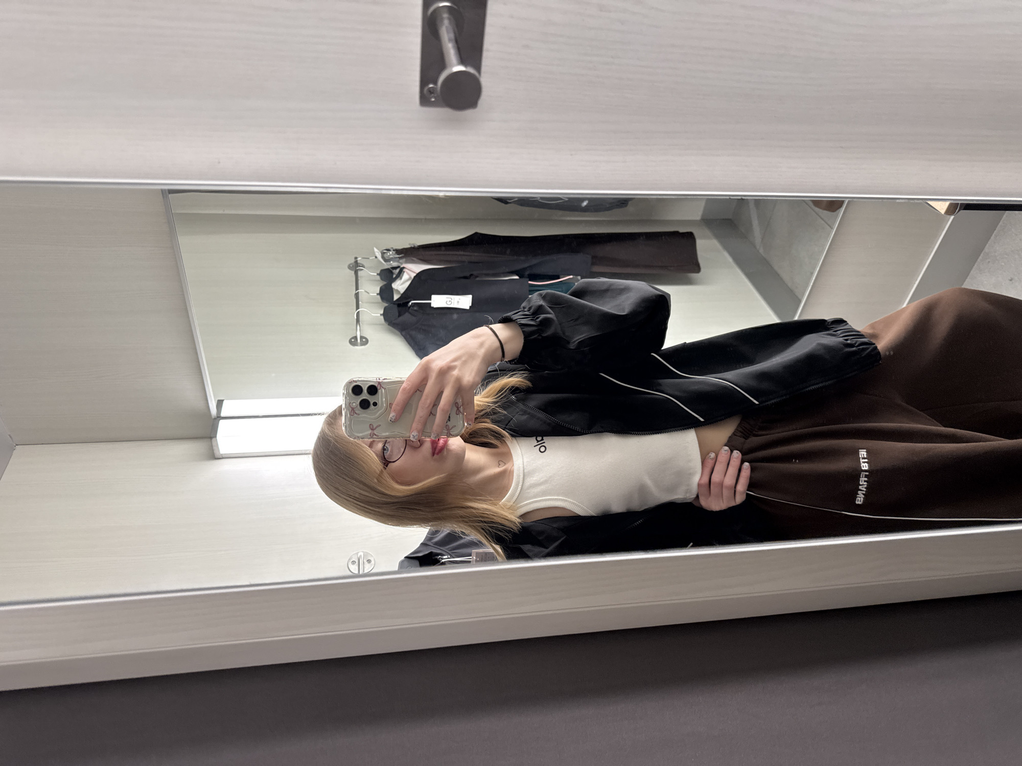 An indoor scene depicts a female individual taking a mirror selfie within a retail changing room in Rostov-on-Don, Russia. The woman, appearing to be a young adult, has shoulder-length blonde hair, wears round-frame glasses with red rims, and a small heart tattoo on her lower neck. She is dressed in a white ribbed crop top with "alo" in black script, a black athletic jacket with white piping, and dark brown sweatpants featuring the reversed text "PETE FRANS" in white on the left thigh. She holds a silver smartphone with a clear, pink-bow-patterned case in her left hand, positioned to capture her reflection. Her right hand rests on her left hip, partially obscured by her jacket. The changing room interior consists of light-colored, possibly paneled, walls, with a darker grey wall section on the left. A linear fluorescent light fixture is reflected above the individual. Behind the woman's reflection, a metal clothing rack holds several dark garments on hangers, some displaying price tags, one of which includes a "GJ" logo. A metallic circular door handle or hook is visible on the right edge of the mirror's frame. The floor reflected beneath the individual appears light-colored.