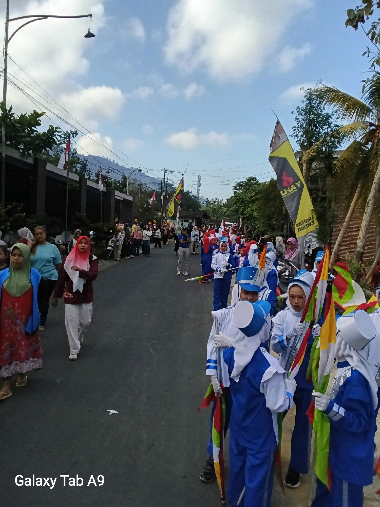 In Bendo, Indonesia, a street is lined with participants in a parade or procession. In the foreground, a group of children in blue and white uniforms, some wearing white headscarves and gloves, hold various colorful flags. Behind them, adults and children are also seen walking, some in traditional attire including headscarves. The street is paved, and there are buildings and trees lining one side. A tall lamppost with a hanging light fixture is visible on the left, and power lines stretch across the sky. Several flags, including the Indonesian flag, are displayed along the street. The sky is partly cloudy with a clear blue expanse. The scene appears to be a daytime event, likely a cultural celebration or holiday.