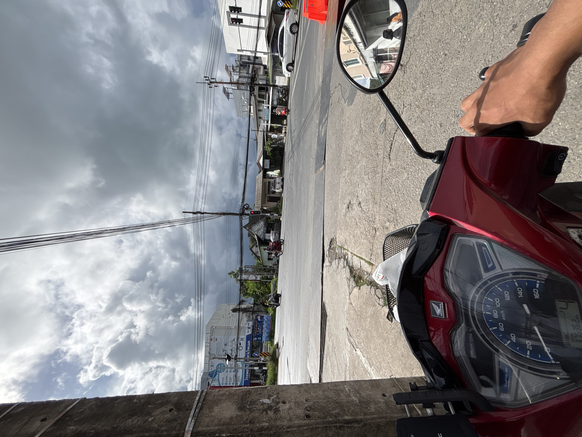The image is a first-person perspective from a red Honda motorcycle parked at a street intersection in Rawai, Thailand. The sky is overcast with dark clouds, suggesting impending rain. In the foreground, the motorcycle's dashboard is visible, showing a speedometer reading approximately 4 km/h and an odometer reading 37536.2 km. A white plastic bag is in the basket in front of the seat.
The intersection features multiple lanes of asphalt road, marked by white lines. Several vehicles are present: a white sedan is stopped at the intersection on the right, a red scooter with two occupants is moving across the intersection from the left, and another motorcycle is seen further back. Buildings line the sides of the road, with a mix of commercial establishments and residential structures. Overhead, a network of electrical wires and poles crisscrosses the sky. The overall impression is of a typical street scene in a tropical urban environment.