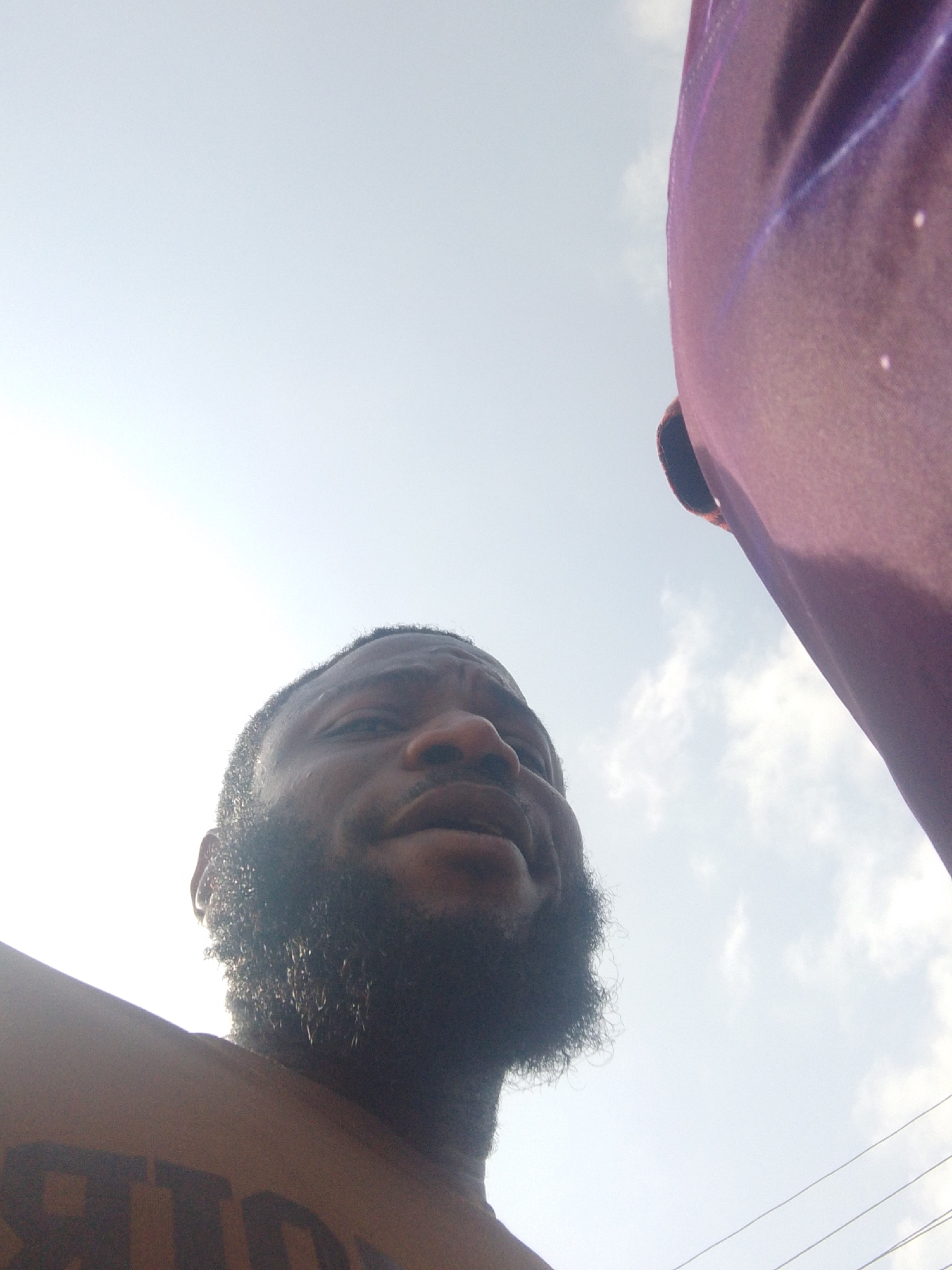 A low-angle photograph shows a man with a full beard looking upward towards a bright, overcast sky. A portion of a purple fabric covers the upper right corner of the image. The man appears to be shirtless, with the word "STRONG" partially visible in black letters on his chest. Based on the prompt, the location is likely Lokoja, Nigeria, although there are no specific details in the image to confirm this. The presence of electric lines in the bottom right corner suggests an urban environment.