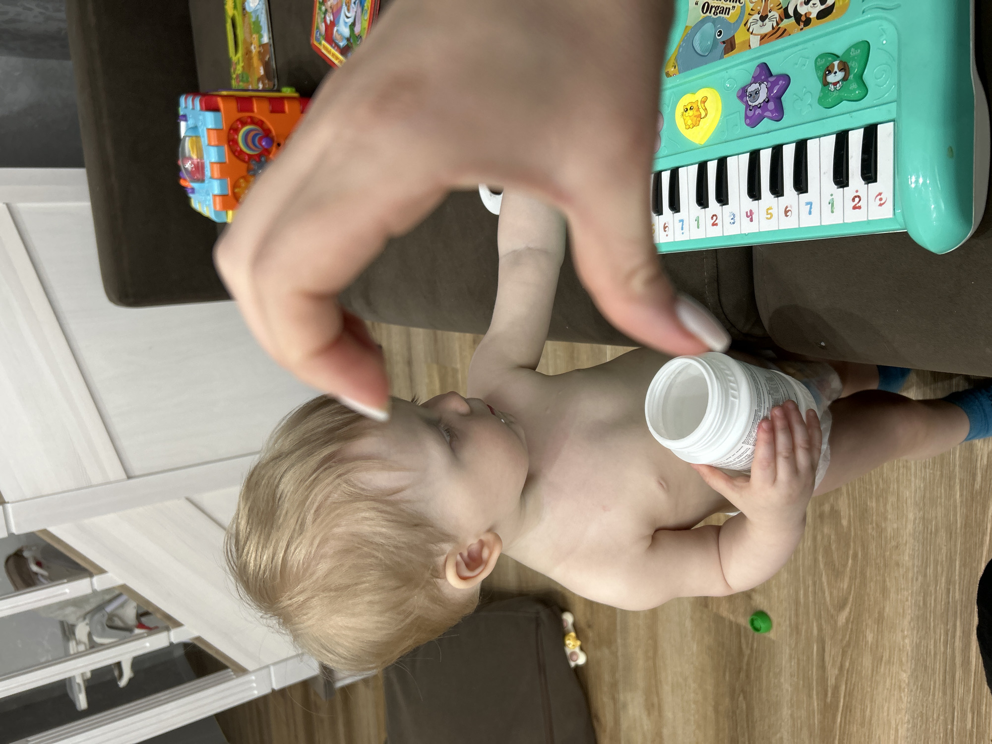 A partially nude toddler is holding a white, cylindrical container. A hand is positioned above the child, as if reaching for them or the container. A toy keyboard and a colorful cube toy are visible in the background, resting on a brown surface. The flooring appears to be made of wood. The location, Chaikovsky, Russia, cannot be determined from the image.