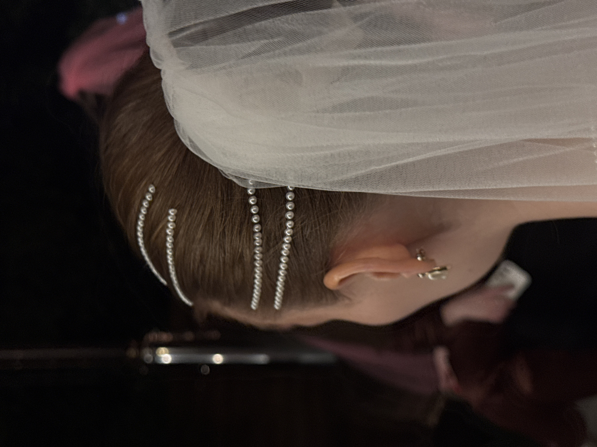 The image shows a close-up of a person's head, tilted downwards. They have brown hair decorated with strands of small, shiny beads or gems, and a white, sheer veil is draped over their head. Part of an ear with a stud earring is visible. The person's skin tone is fair, and part of their cheek and neck can be seen. The background includes a dark, reflective surface, which seems to be a piano or a similar glossy object, and a blurry dark pink shape. The location and city are not identifiable from the image.