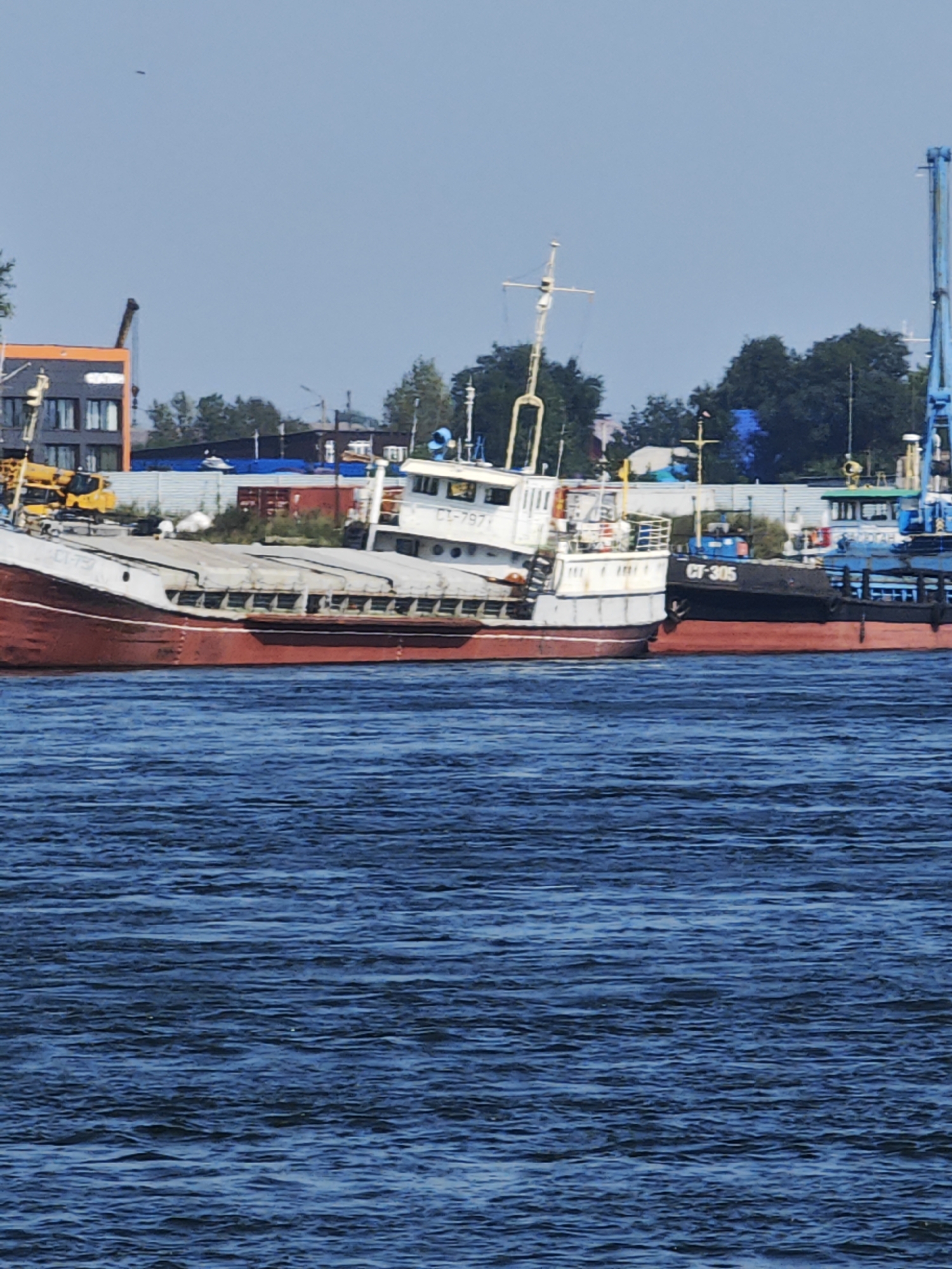 In Krasnoyarsk, Russia, two docked ships are visible on a body of water. The larger ship, on the left, is white with a red hull. It has a long, covered deck. On its side, the number "CT-797" is visible. To the right of this ship is a smaller, black ship with a red hull. The number "CT-305" is painted on its side. The background shows industrial buildings, trees, and cranes, indicating a port or shipyard. The sky is clear and blue. The water is choppy and deep blue.