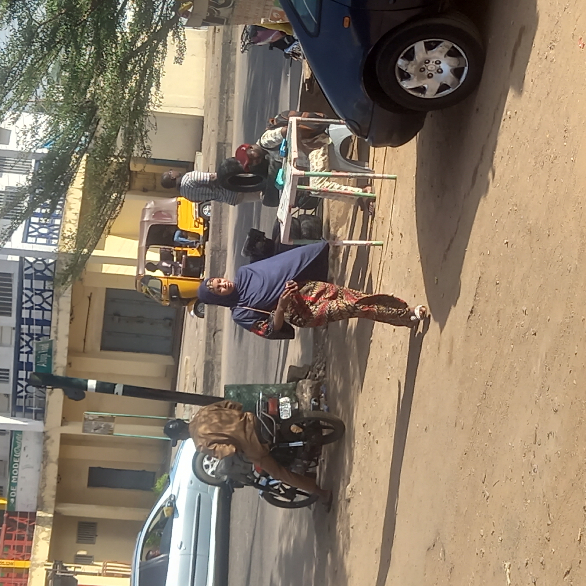 The image depicts a street scene in Dadi Riba, Nigeria. A woman wearing a blue headscarf, navy blouse, and patterned skirt is walking on the sidewalk. A man sits on a motorcycle parked nearby. A blue car is also parked close to the edge of the sidewalk. Across the street, there is a yellow motorized tricycle vehicle and a vendor with a makeshift table, attended by two men, appears to be selling items, possibly tires. A building with a sign "DE MODEL Outfit" is also visible in the background. The environment appears to be a busy urban street with a sandy colored road and sidewalk.