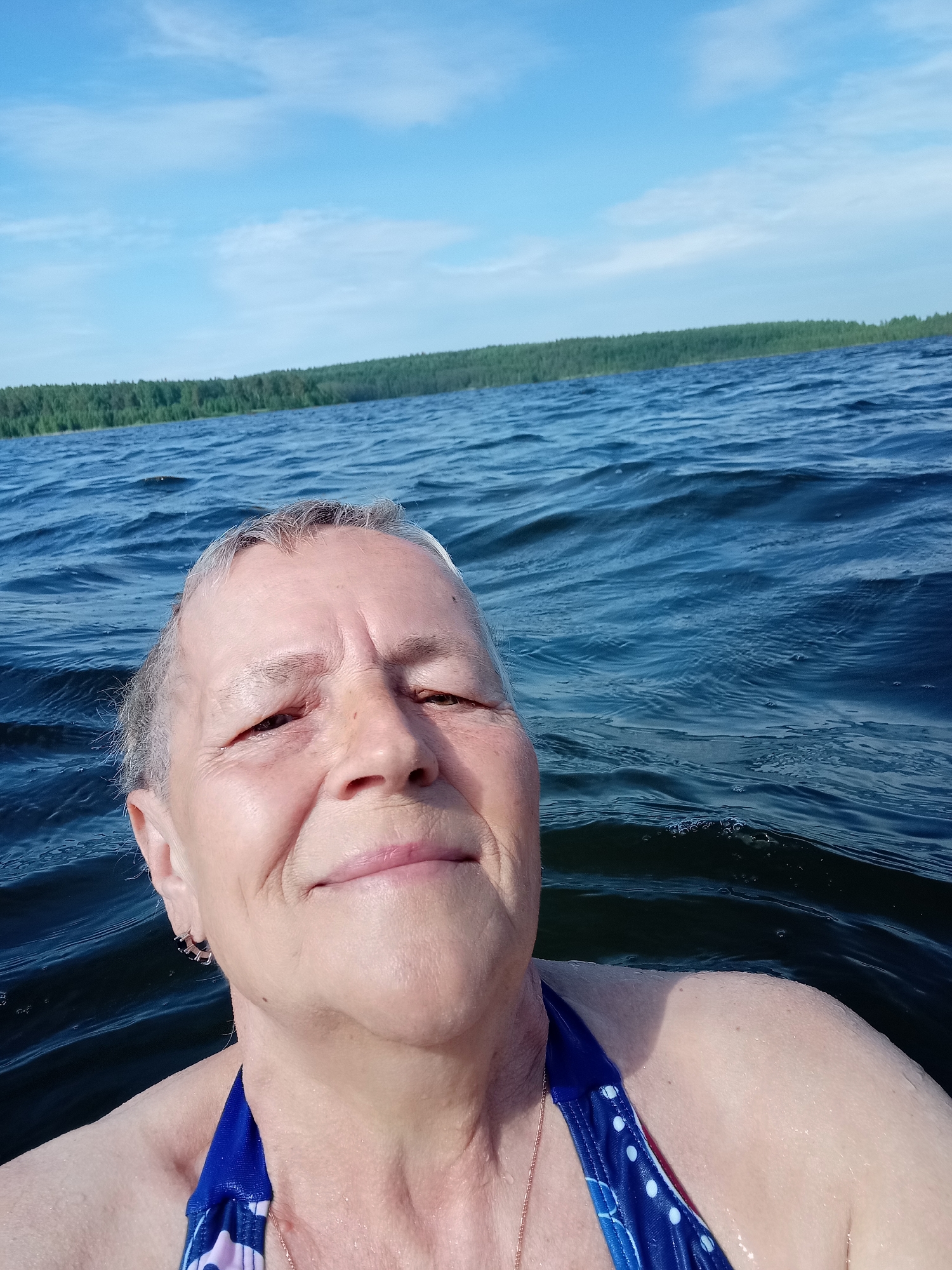 A mature woman is partially submerged in a body of water, likely a lake, with her head and upper torso visible. She is wearing a blue and white patterned swimsuit. The water is dark blue and slightly choppy with small waves. In the background, there is a line of green trees along the horizon, suggesting a shoreline. The sky is blue with some scattered white clouds. The image appears to be a selfie taken by the woman. The location is potentially the city of Degtyarsk, Russia, where the lake is a typical landscape.
