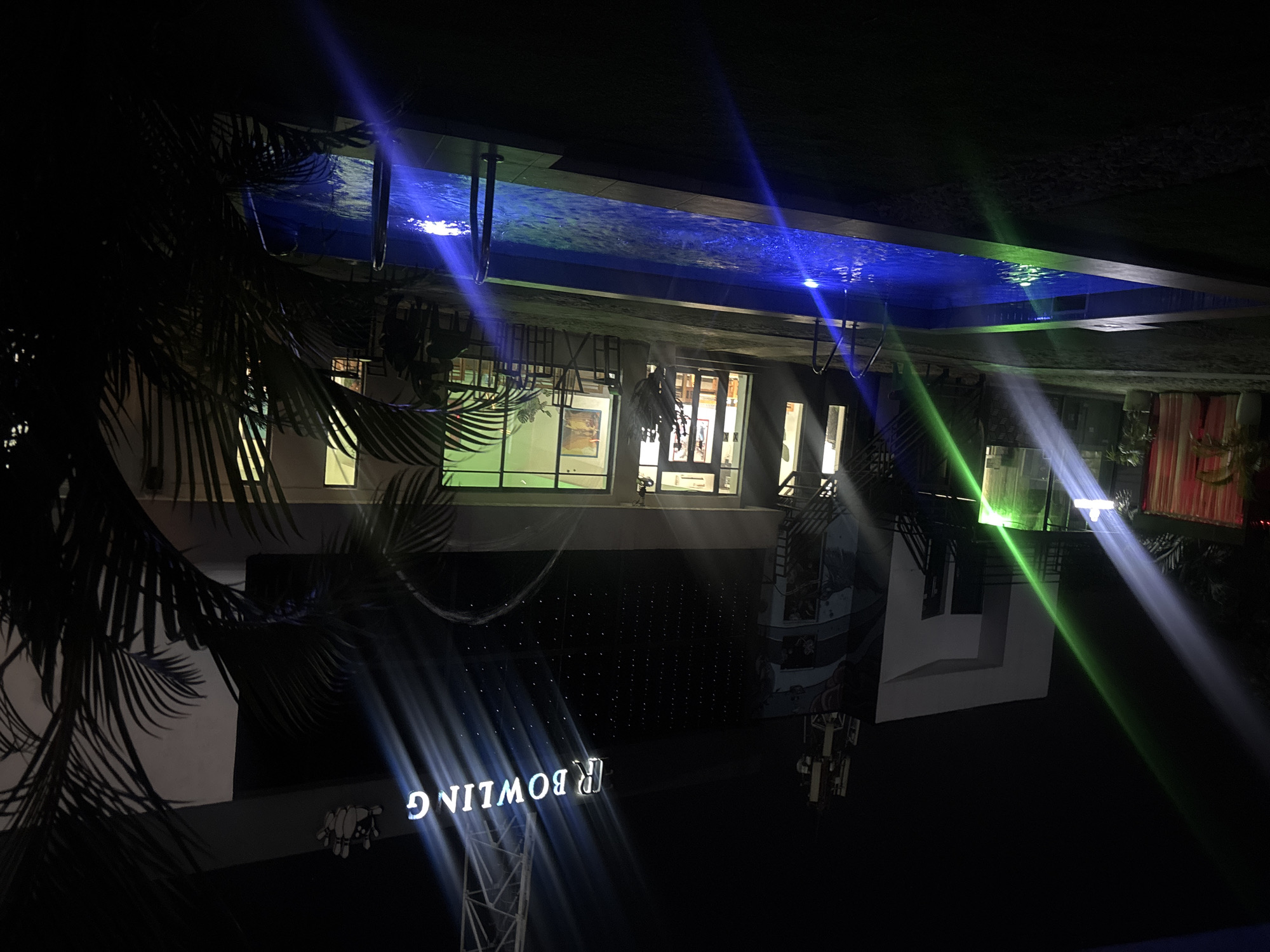 The image shows an evening scene in Port Harcourt, Nigeria. A building is visible with a brightly lit "BOWLING" sign. The building also features a rooftop swimming pool illuminated with blue lighting. Palm trees are partially visible on the left side of the frame. The lighting on the building suggests it is a recreational or entertainment venue.
