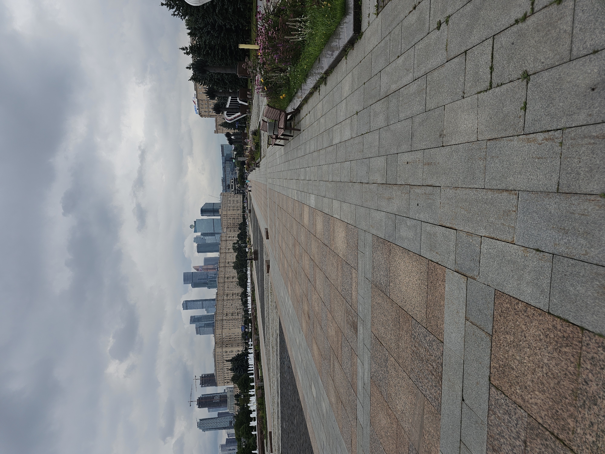 The image depicts a wide, paved pedestrian walkway in Moscow, Russia, under an overcast sky. The walkway is constructed from large granite tiles, with sections of different colored and textured granite. To the left of the walkway, a large open area with patterned paving stones is visible, leading towards a series of fountains in the distance. Beyond the fountains, a long, low-rise building with a facade of numerous windows stretches across the background.
Dominating the skyline behind this building is a collection of modern skyscrapers, forming a cityscape characteristic of Moscow's business district, likely Moscow City. The skyscrapers vary in height and design, with some appearing to be under construction, indicated by cranes. To the right of the walkway, a strip of greenery with flowering plants and trees lines the path, interspersed with several dark-colored park benches. Further to the right, a dense cluster of evergreen trees stands, partially obscuring another building in the distance. No people are clearly visible in the immediate foreground, but a few distant figures can be observed walking along the paved area. The overall scene conveys an urban environment with both historical and contemporary architectural elements.