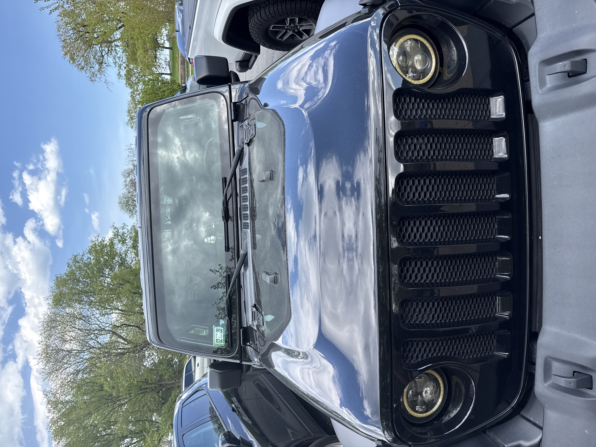 A close-up, slightly canted shot focuses on the front end of a black Jeep Wrangler, parked in an outdoor lot in Chicago, Illinois. The vehicle's hood reflects the bright blue sky filled with scattered white clouds. Visible are the headlights with halo rings, the seven-slot grille, and windshield with windshield wipers. A green sticker is visible on the lower right corner of the windshield. Adjacent to the black Jeep, the corner of a white truck is partially visible, with a dark tire and wheel. In the background, trees with green foliage are discernible.