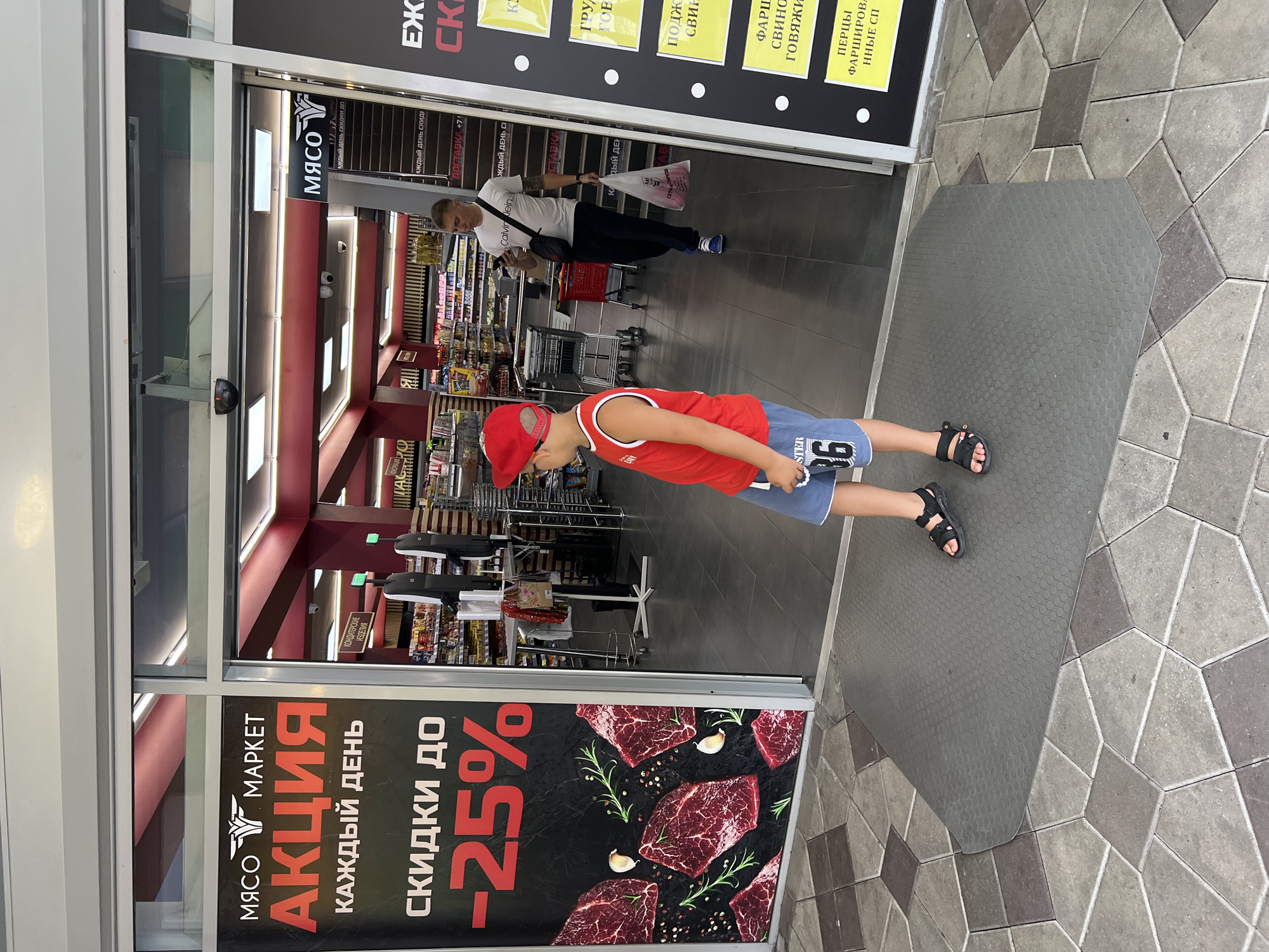 This is a medium shot, eye-level, outdoor photograph taken in front of a supermarket entrance. The photo features a young boy in the foreground, facing away from the camera and looking into the store. He is wearing a red sleeveless shirt with a white trim, blue shorts with the number "36" printed on them, and black sandals. He is standing on a grey mat at the entrance. To the right, a man in a white t-shirt with "Calvin Klein" printed on it, dark pants, and blue sneakers is walking into the store while looking at his phone and carrying a plastic bag with red items inside. The supermarket entrance has glass doors and a large black banner on the left side displaying text in red and white. The text on the banner includes "МЯСО МАРКЕТ" (Meat Market) and "АКЦИЯ КАЖДЫЙ ДЕНЬ" (Action Every Day) with a discount of "СКИДКИ ДО -25%" (Discounts up to -25%). The interior of the store is visible, showing aisles stocked with products and illuminated by overhead lighting. The pavement outside the entrance is made of grey and brown geometric tiles. The overall scene suggests a commercial setting, likely a meat market or grocery store, with promotional signage visible. The location is identified as Sochi, Russia.