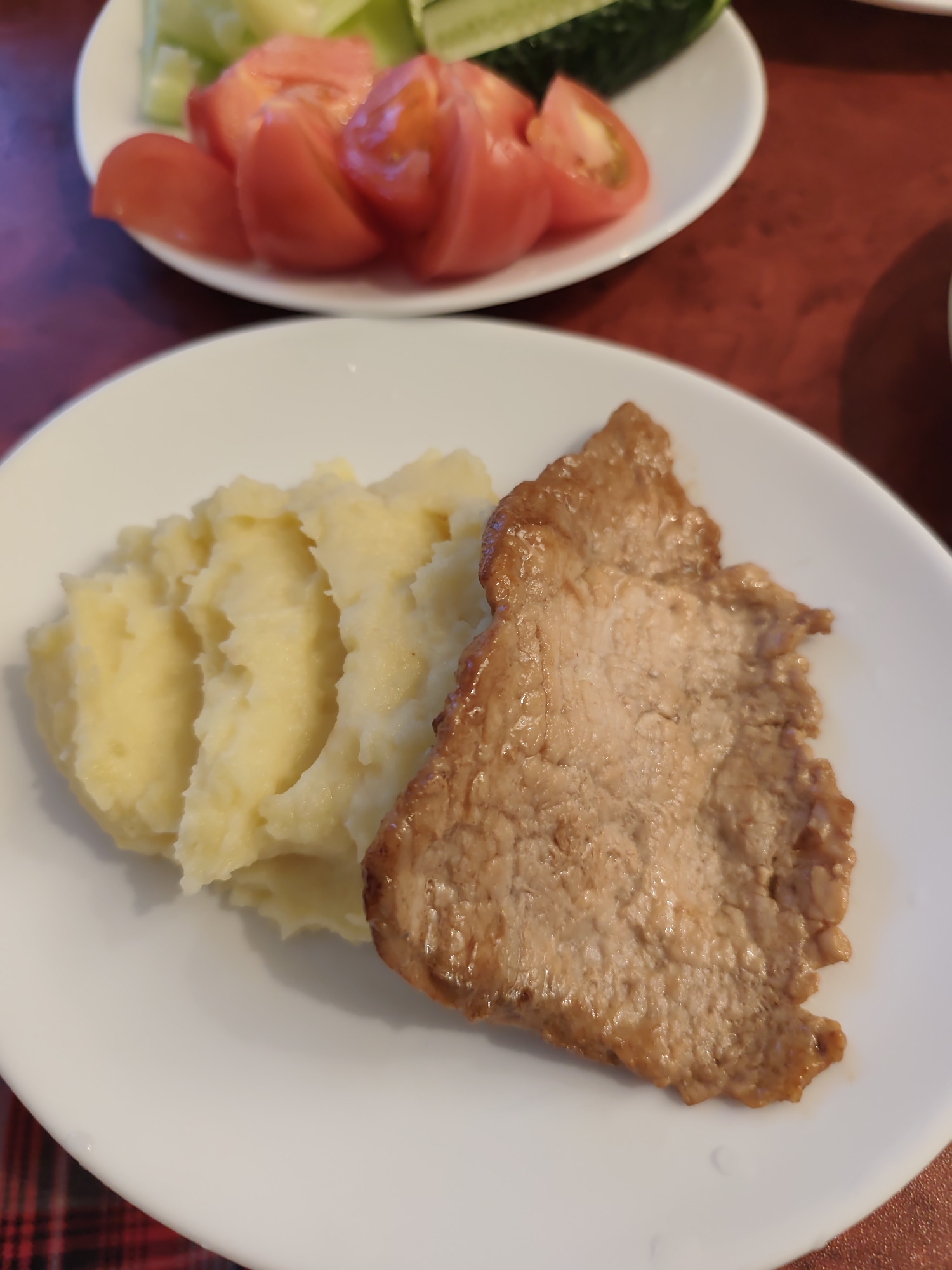 A close-up, elevated view captures a plated meal indoors. In the foreground, a white, round ceramic plate holds a serving of mashed potatoes on the left and a single piece of cooked meat on the right. The mashed potatoes are light yellow, textured with soft peaks and indentations. The meat is an irregularly shaped, light brown cutlet, appearing seared or roasted, with a slightly glossy surface. In the background, out of focus, another white plate is partially visible, containing red sliced tomatoes and green cucumber slices. The plates rest on a reddish-brown table surface, possibly wood or laminate, with a hint of a red and black plaid pattern visible in the bottom left corner, likely a placemat or tablecloth. No individuals are present.