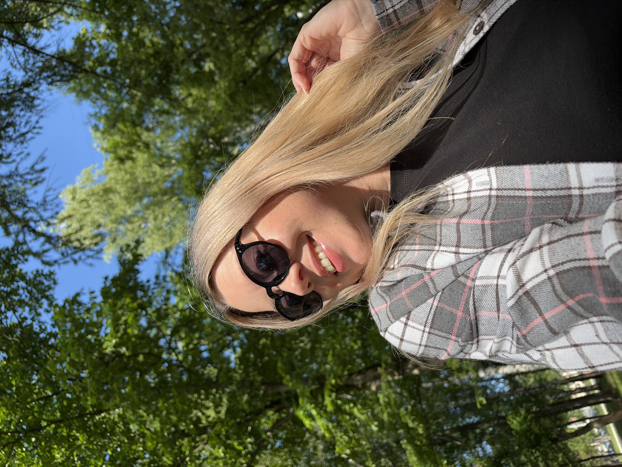 An eye-level portrait captures a young woman outdoors, looking upwards with a bright smile. She has long, straight blonde hair, some of which she is gently holding with her right hand, positioned above her head. She wears black, rounded sunglasses and a black top, layered with a grey, white, and pink plaid shirt that features thin dark grey lines. The shirt is unbuttoned at the collar. Her smile is broad, revealing white teeth, and her lips appear glossy and pink.
The setting is outdoors, likely a park or a natural green space in Chaikovsky, Russia, as suggested by the lush green foliage and clear blue sky visible in the background. The photograph is taken from a low angle, emphasizing the overhead canopy of bright green deciduous trees against the vibrant blue sky, indicating bright daylight and clear, sunny weather. The strong natural light illuminates her hair and face. The overall mood conveyed is cheerful and relaxed, suggesting she is enjoying a pleasant day outdoors. There is no visible text in the image.