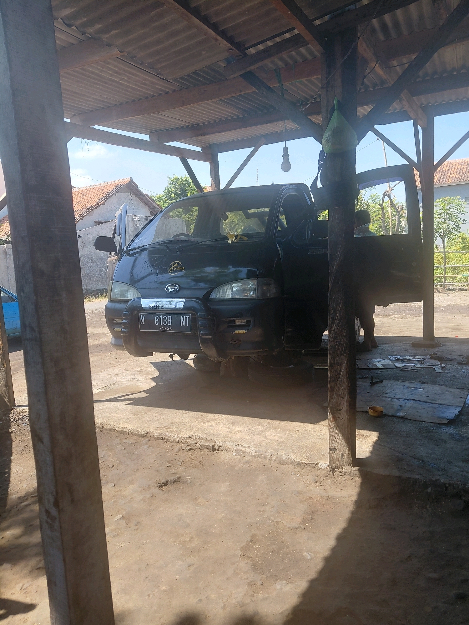 A black van is parked under a corrugated tin roof supported by wooden beams and posts. The van's front license plate reads "N 8138 NT 11-26". The driver's side door of the van is open, and a person, partially obscured, is visible inside. Another person stands near the open door. The ground is dirt and concrete, with some scattered debris. In the background, houses with red-tiled roofs and trees are visible under a clear blue sky. This scene is located in Paiton, Indonesia.