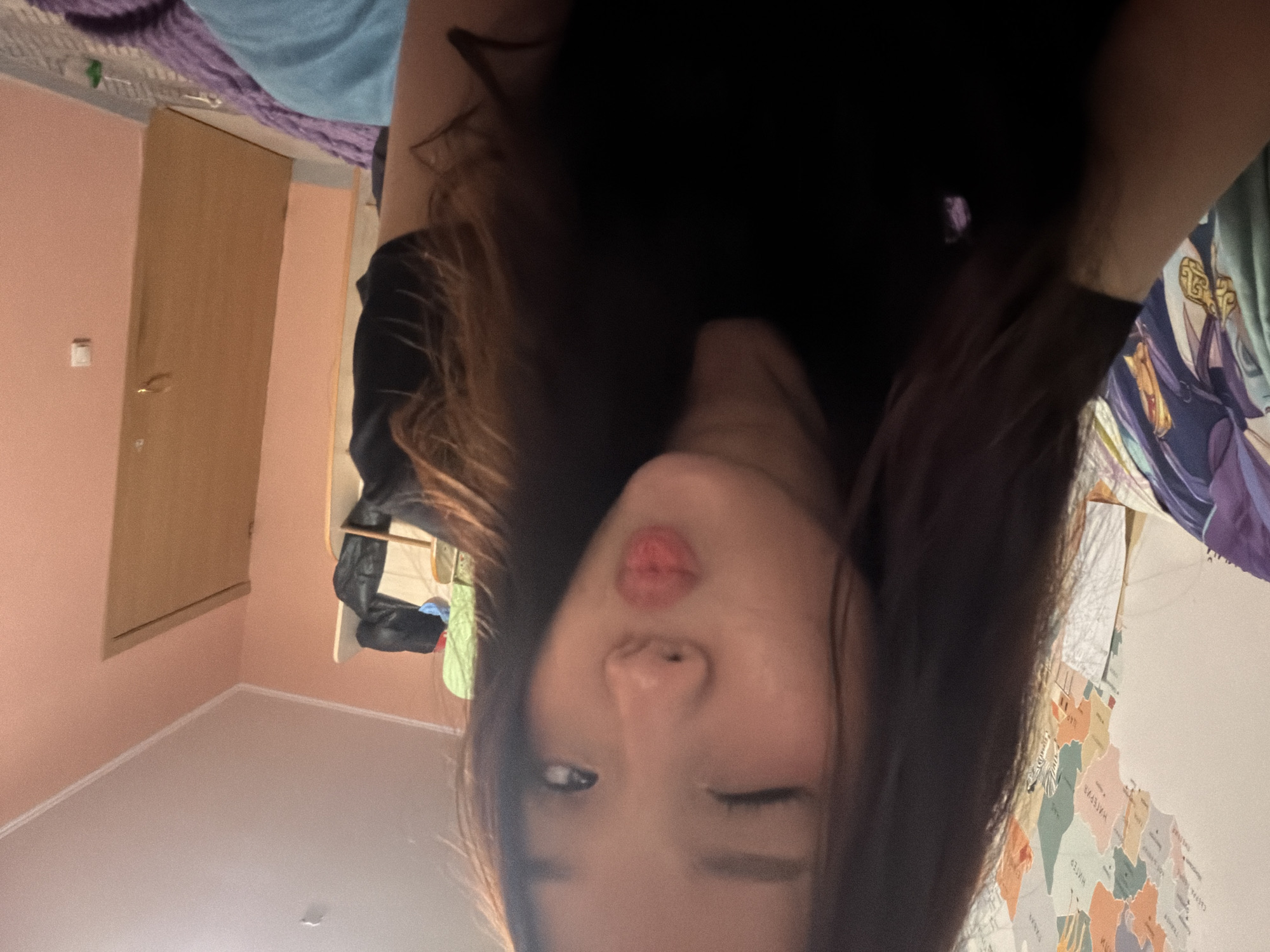The image shows a young woman taking a selfie in a room. She is wearing a black t-shirt and has her hair down. She is winking and puckering her lips, giving a playful expression. In the background, a colorful map of the world is visible on the wall. There is also a closet with some clothes hanging on it, and a door painted in a peachy color. The lighting suggests that the photo was taken indoors, likely in a bedroom. The room appears to be somewhat cluttered, with blankets and other items visible. The overall impression is one of a casual, personal moment captured in an everyday setting.
