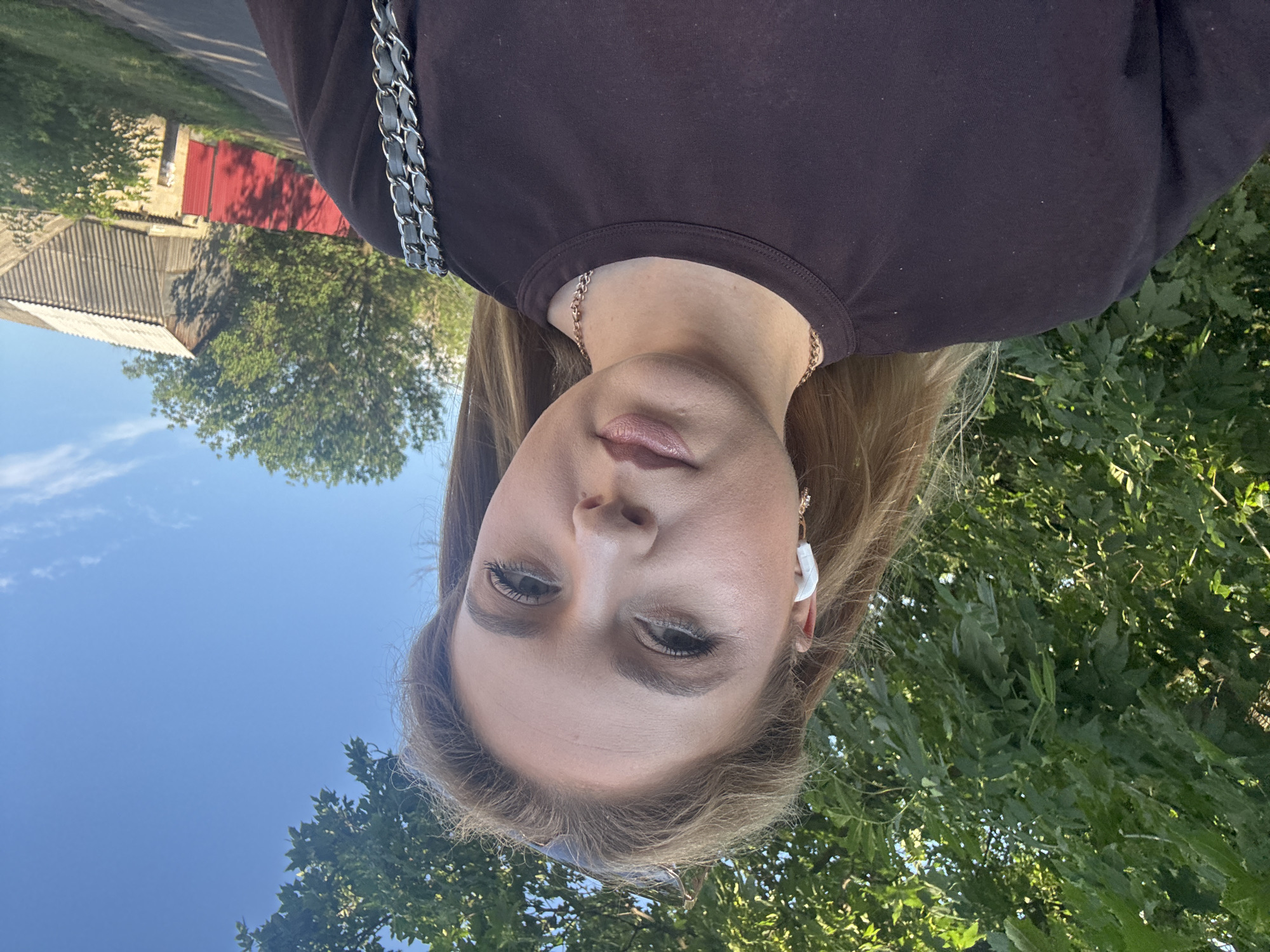 A young woman with blonde hair and fair skin is taking a selfie outdoors. She is wearing a dark brown crewneck t-shirt and has white wireless earbuds in her ears. A delicate gold chain necklace is visible around her neck, and part of a silver chain purse strap is seen over her right shoulder. The background shows lush green trees and a bright blue sky with some scattered clouds. In the distance, a rustic building with a corrugated roof and a red fence are visible. The lighting suggests it is daytime.
