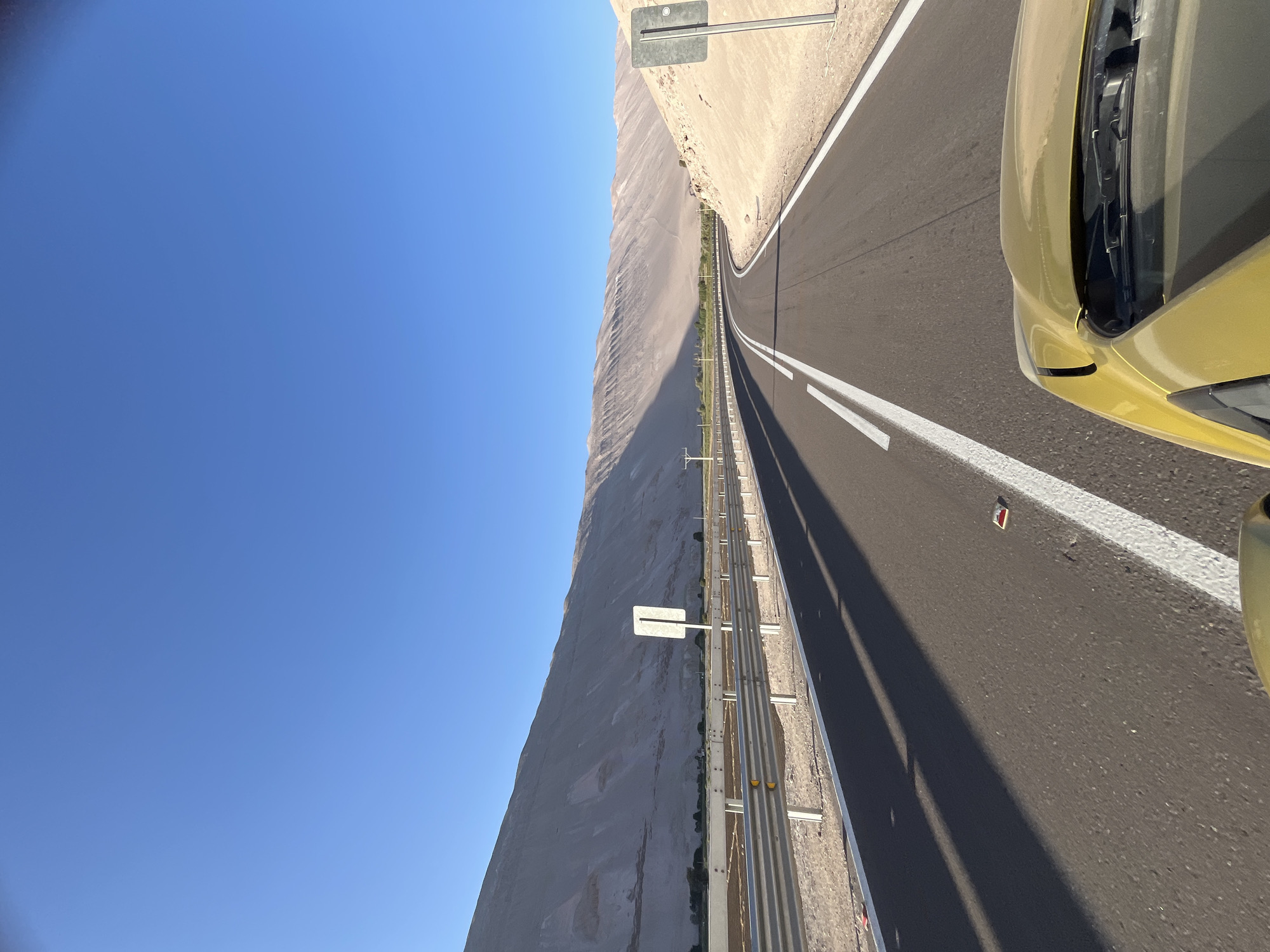 A view from inside a yellow car, driving on a highway in Arica, Chile, heading toward the horizon under a clear blue sky. The road has a white line down the center, and a safety barrier lines the right side of the road. A light-colored mountain range is on the left side of the road. Road signs are visible along the edge of the highway.